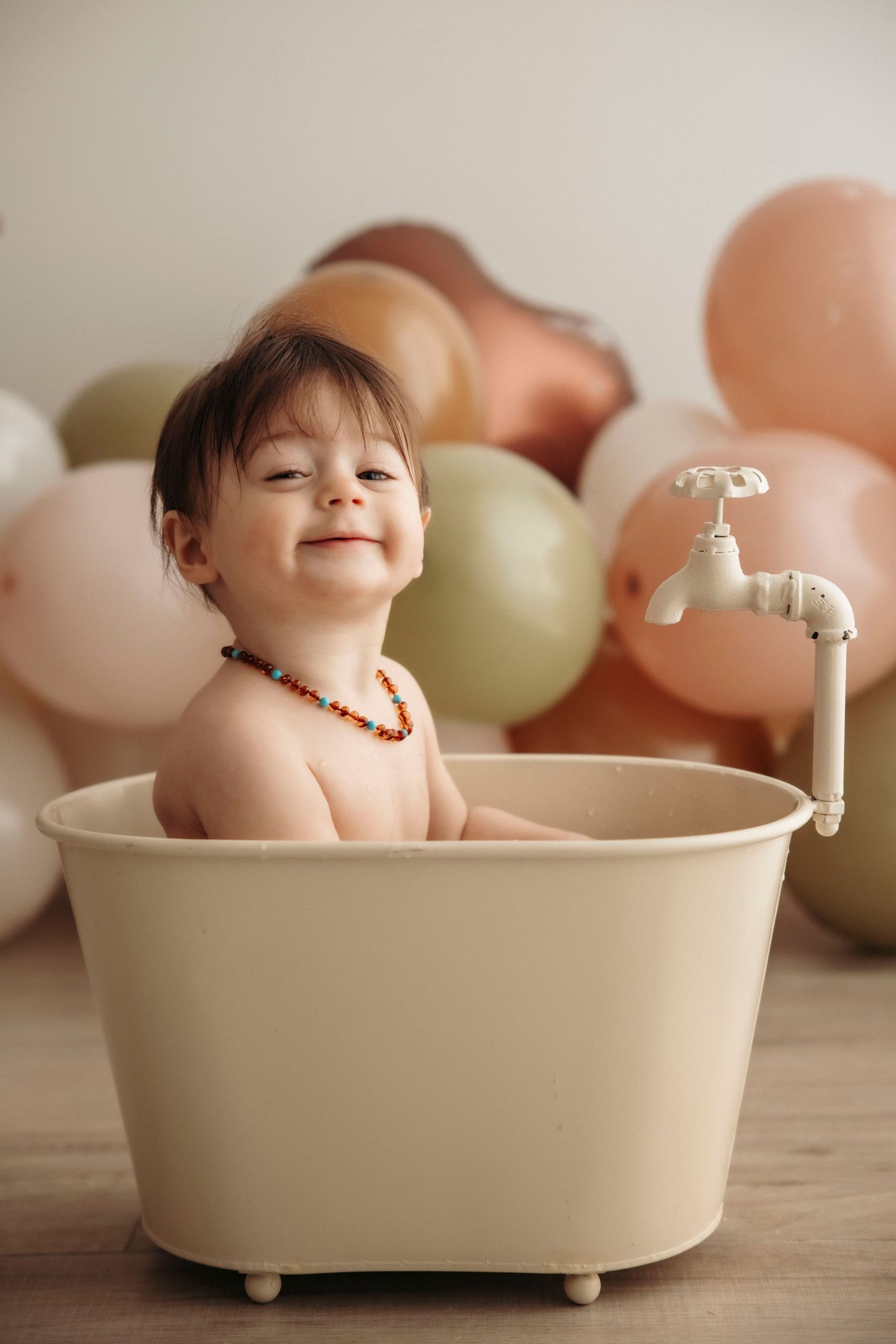 Cute baby sitting in a small vintage white bathtub with a faucet, wearing an amber teething necklace.
