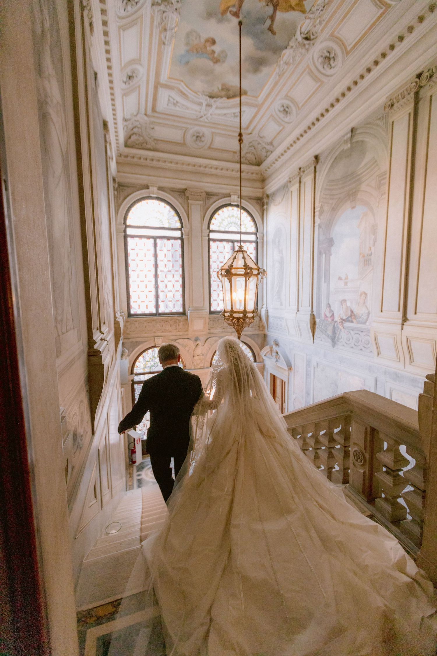 Black and white photo of a bride in a lace gown with veil and groom in tuxedo posing on lawn outside elegant villa.