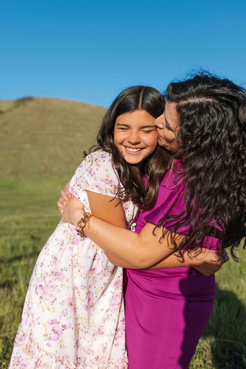 Woman in purple dress kisses smiling girl in floral dress while embracing outdoors on green hillside.