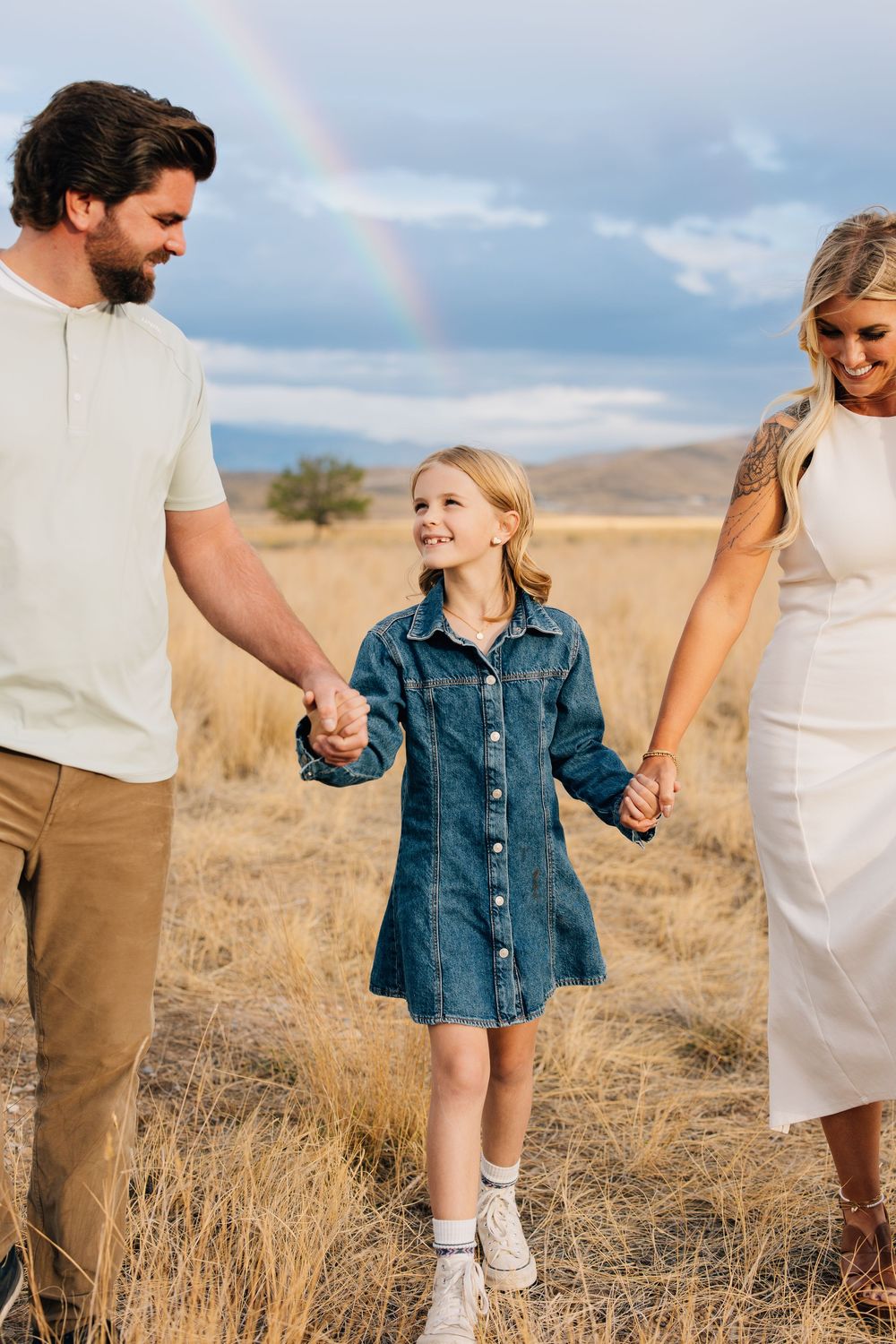 Smiling girl in denim dress holds parents hands in golden field with rainbow in background.