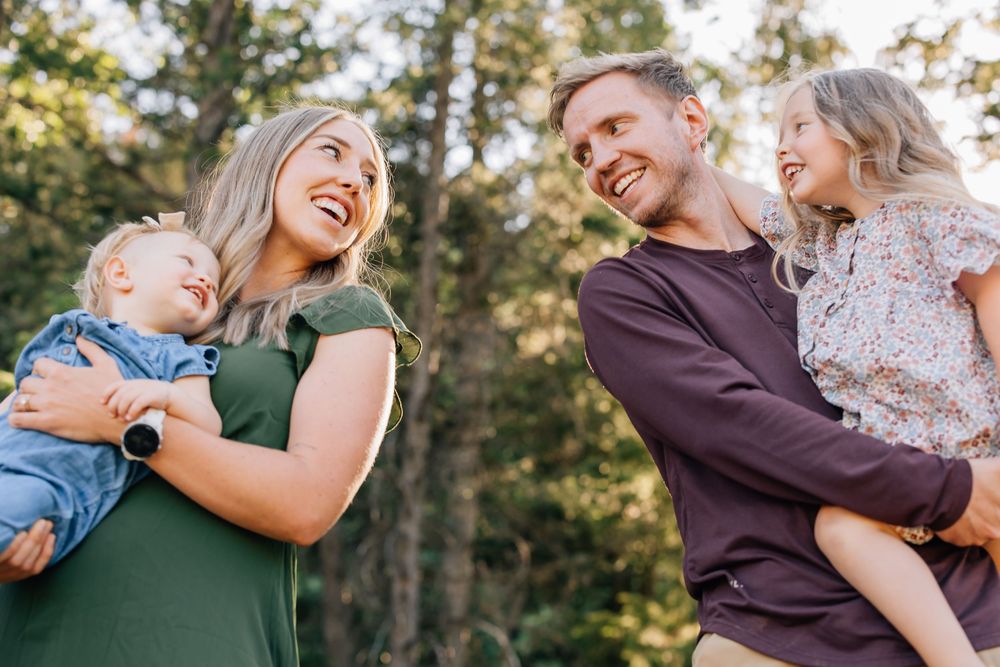 Happy family of four smiling in the Utah outdoors, parents holding two young children with tall green trees in background.