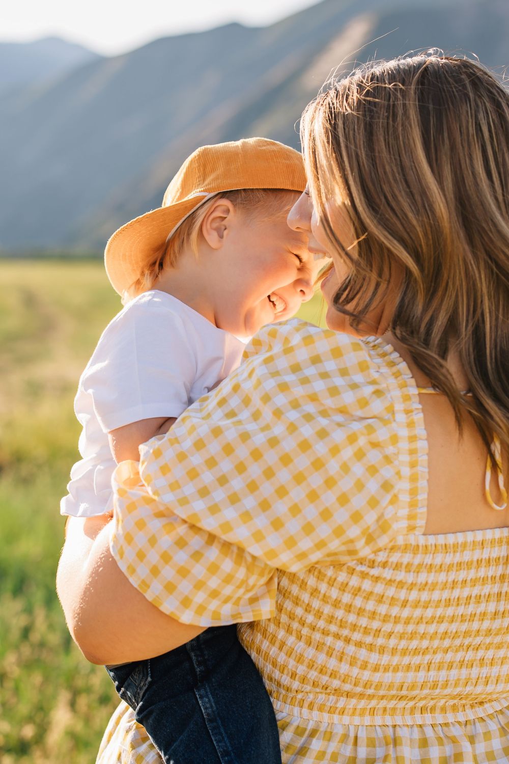 Mother in yellow gingham dress holding laughing toddler in orange hat against mountain backdrop.