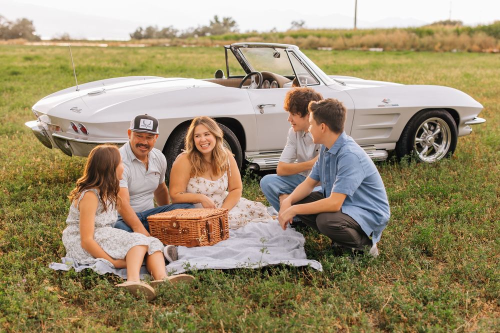 Family of five enjoying a picnic on a blanket beside a classic white Corvette convertible in a green field.