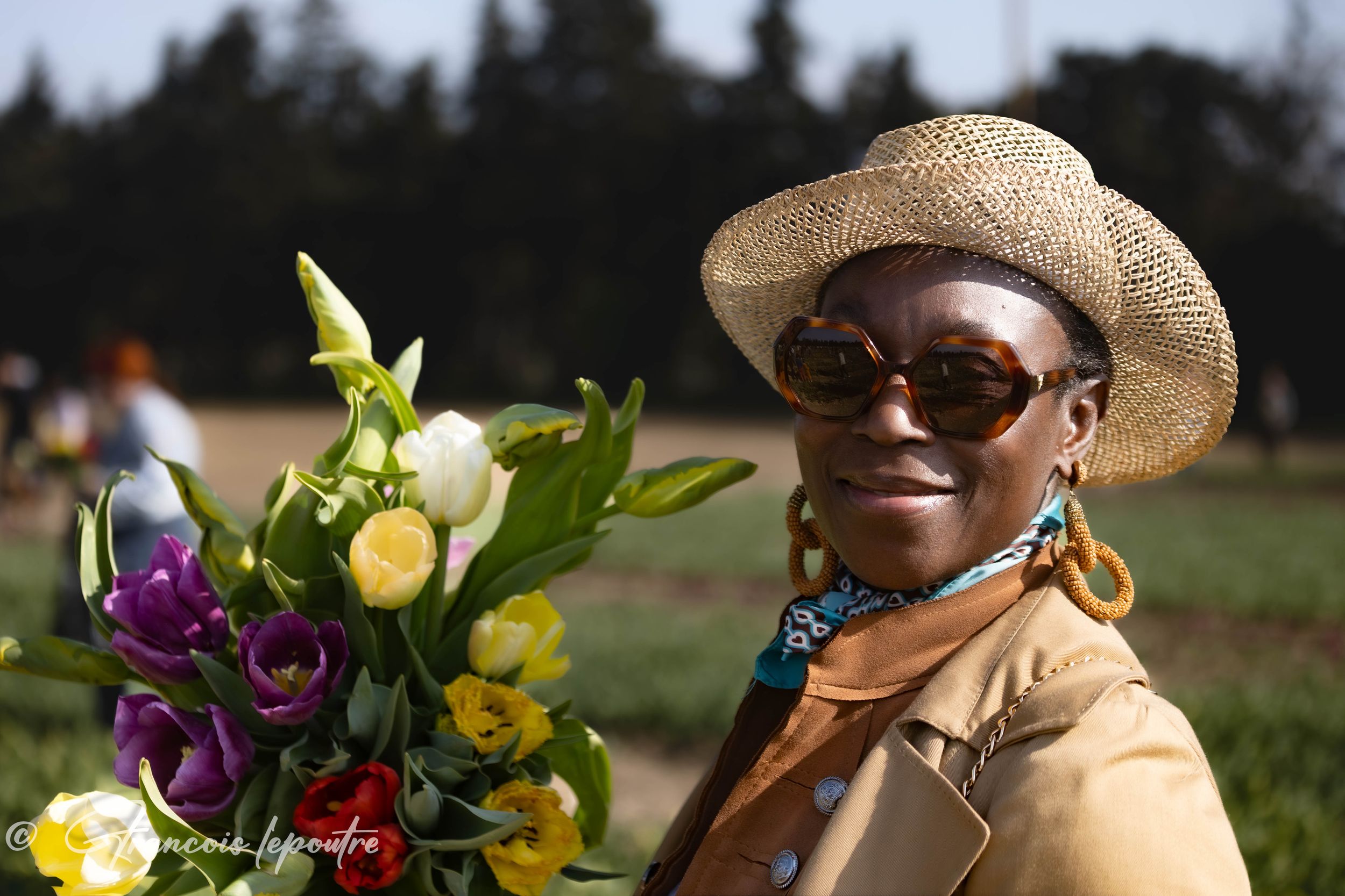 A woman wearing a straw hat and sunglasses holds a colorful bouquet of tulips in an outdoor flower field.