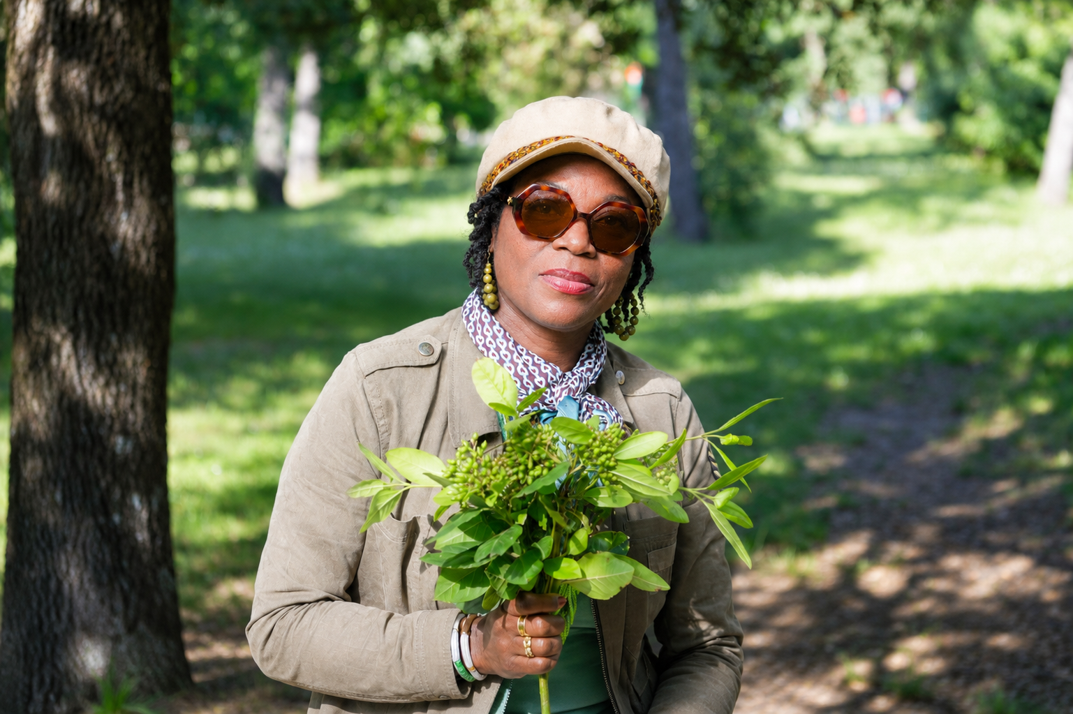 Stylish woman wearing sunglasses and a beret holding a bouquet of fresh green plants in a sunny park.
