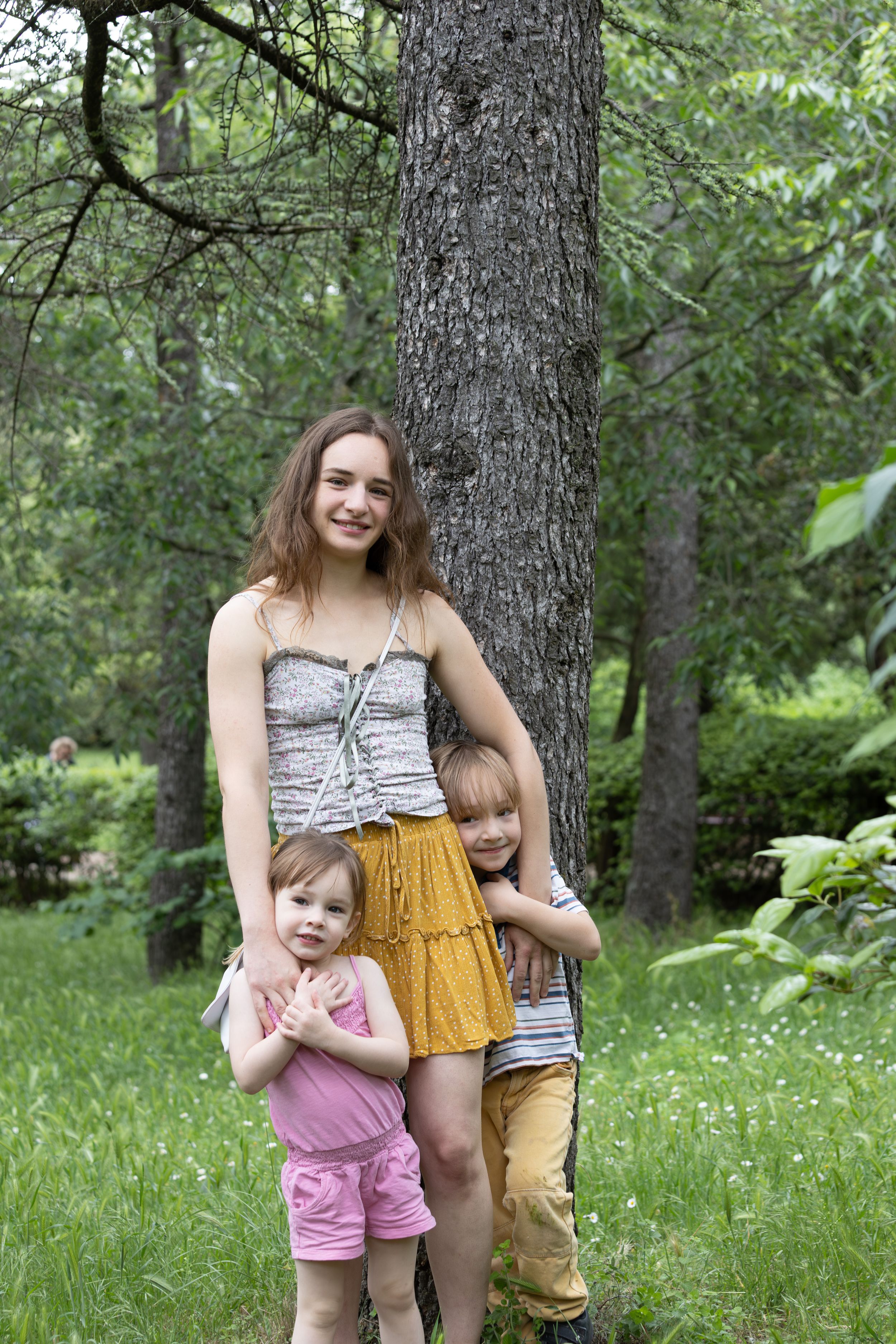 A smiling teenage girl poses with two young children in front of a tree in a green forest.