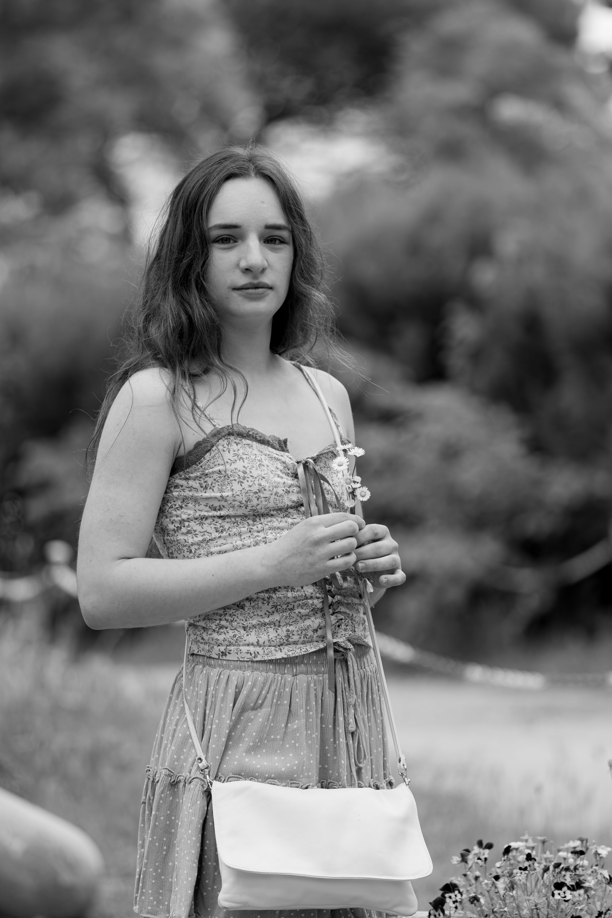 Black and white portrait of a young woman in a summer dress holding flowers in a lush garden setting.