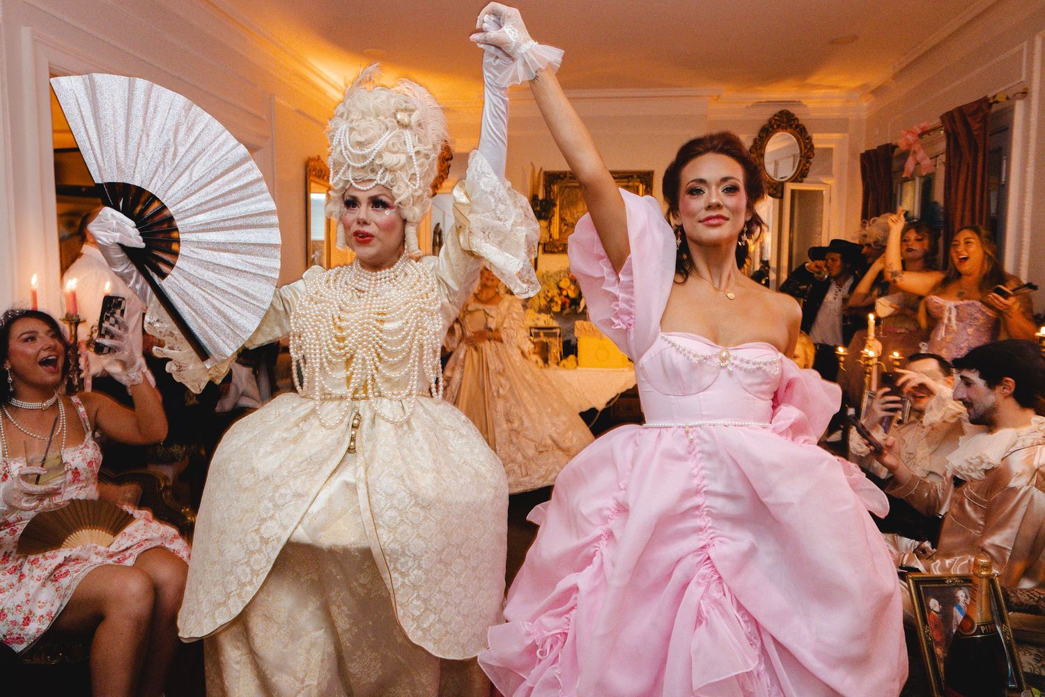 Two women in elaborate Marie Antoinette costumes dance at a glamorous themed party.