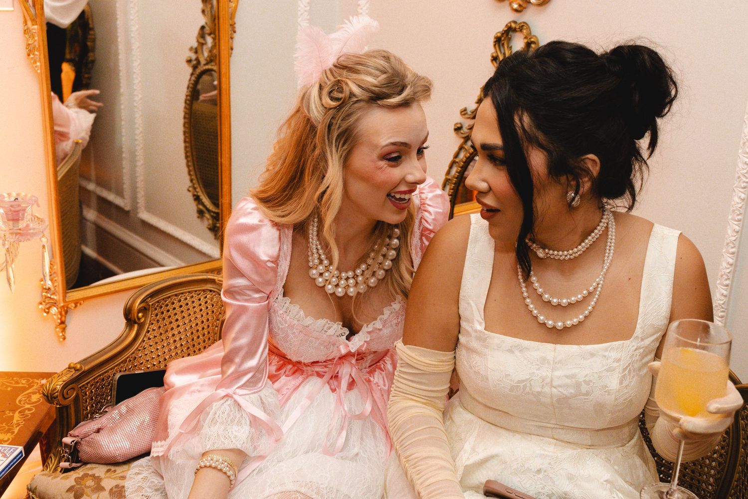 Two women in elegant Marie Antoinette-inspired pink and white gowns with pearl necklaces laughing together at a themed event.