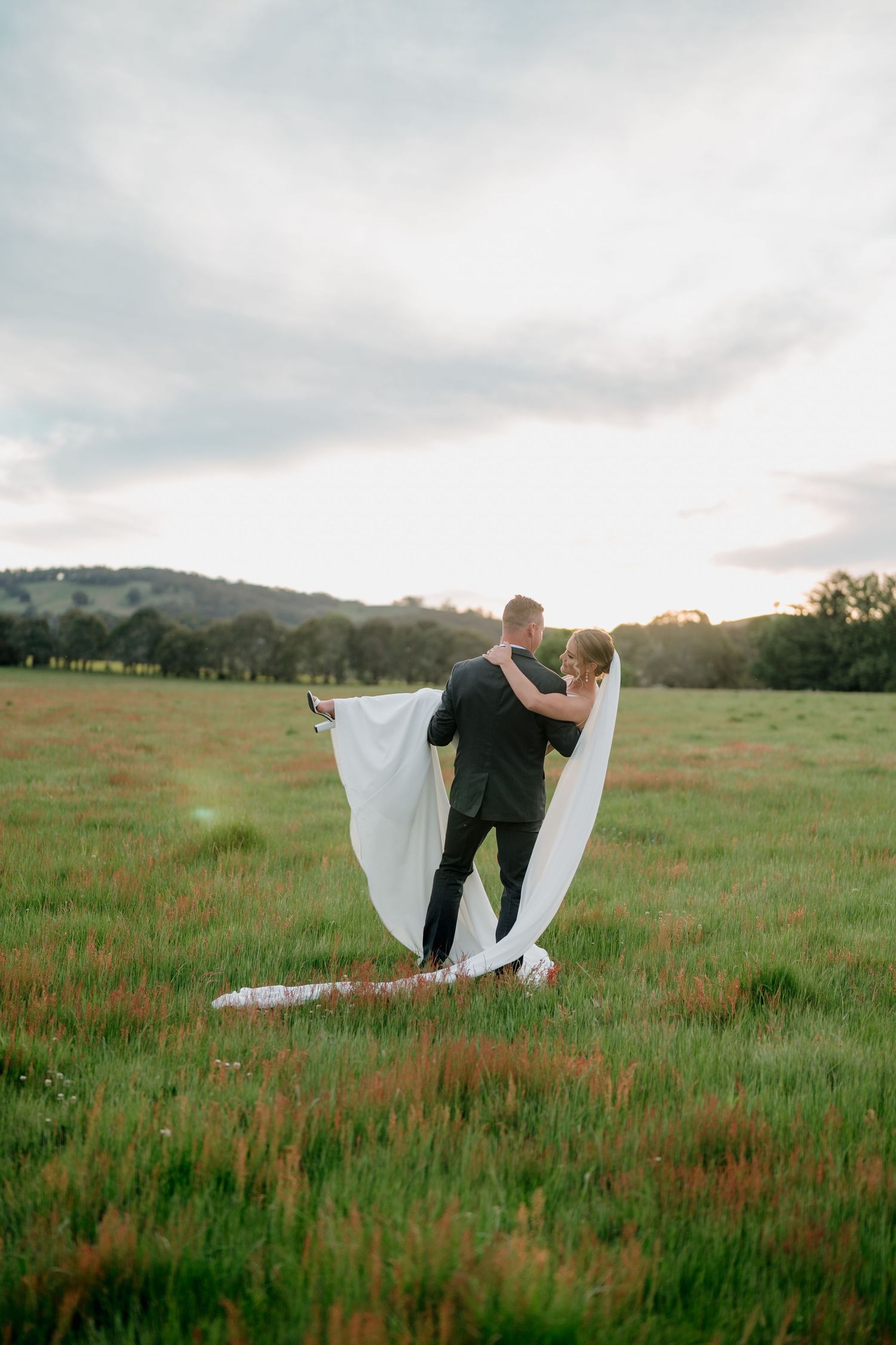 Groom lifts bride in white gown and veil in a lush green field at sunset with rolling hills in the background.