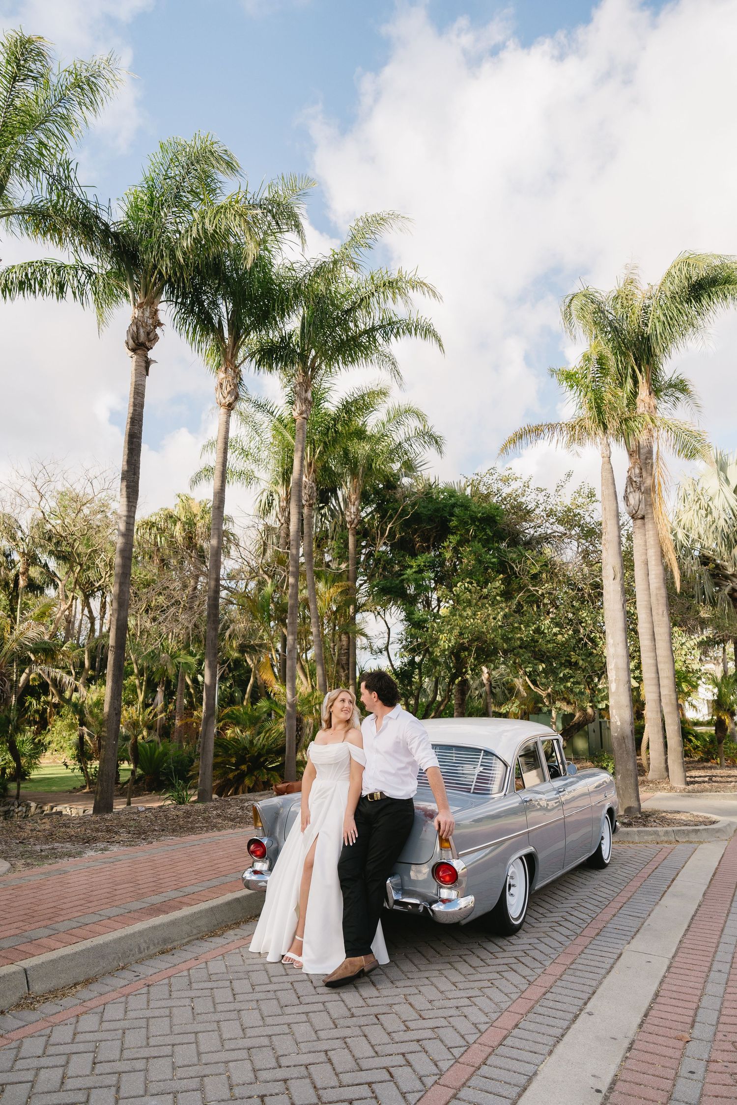 Groom opens vintage gray car door for bride under blooming jacaranda tree on wedding day.