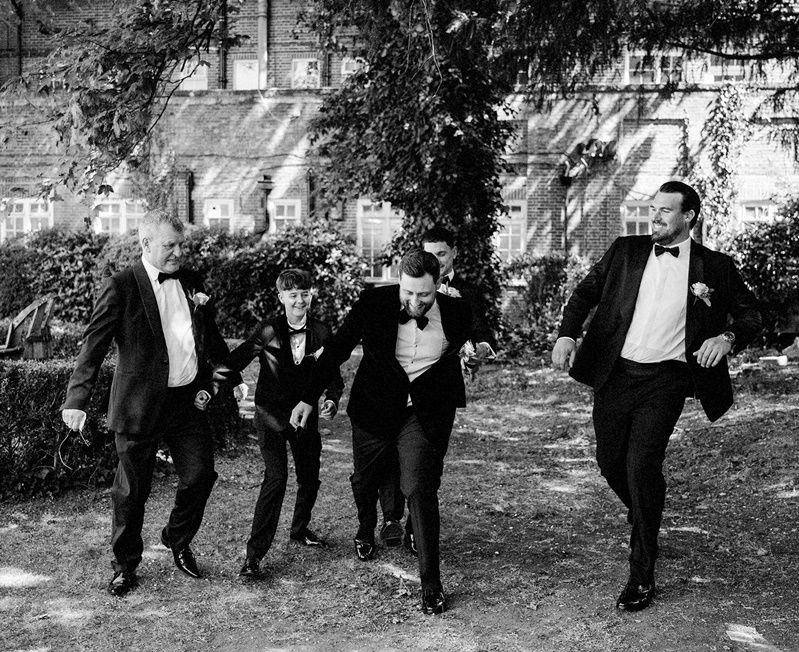 Black and white photo of groomsmen in tuxedos running and laughing outside a brick building on a wedding day.