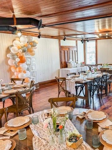 Rustic event venue with wooden tables, cross-back chairs, floral centerpieces, and orange-gray balloon arch decor.