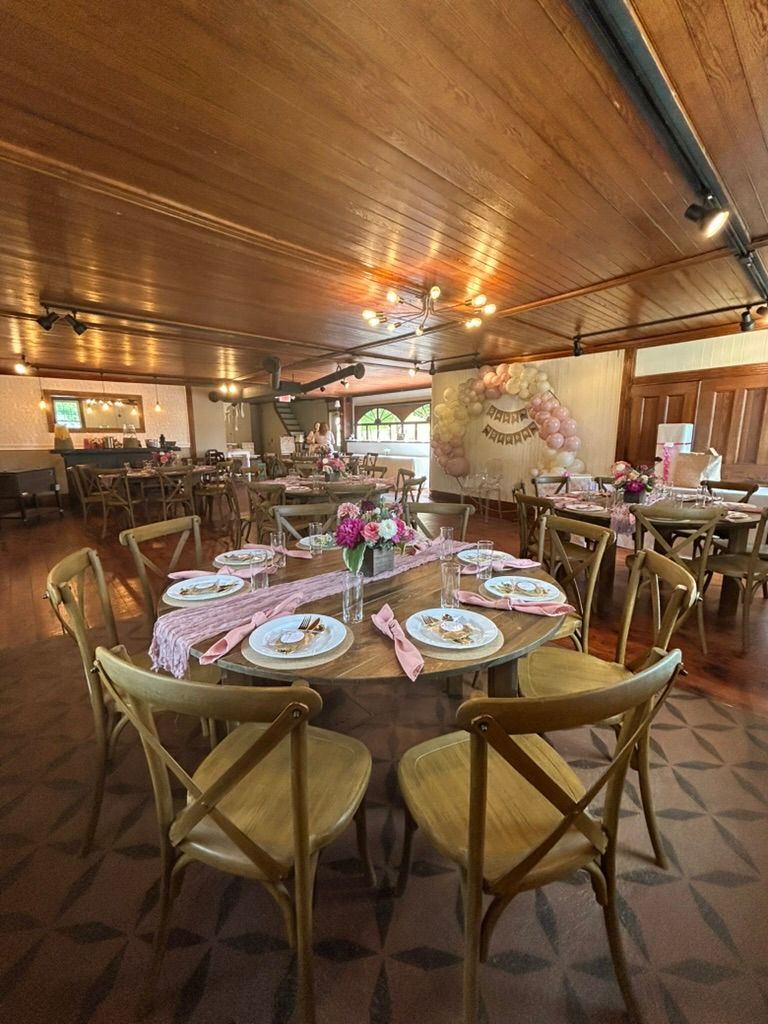 Elegant event venue with round wooden tables, cross-back chairs, pink table settings, and balloon arch backdrop.