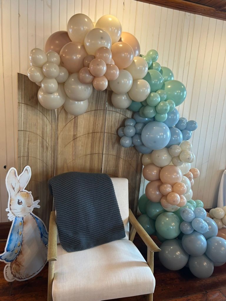 Peter Rabbit themed baby shower balloon arch in pastel blue, green, and tan tones with bunny cutout beside white chair.