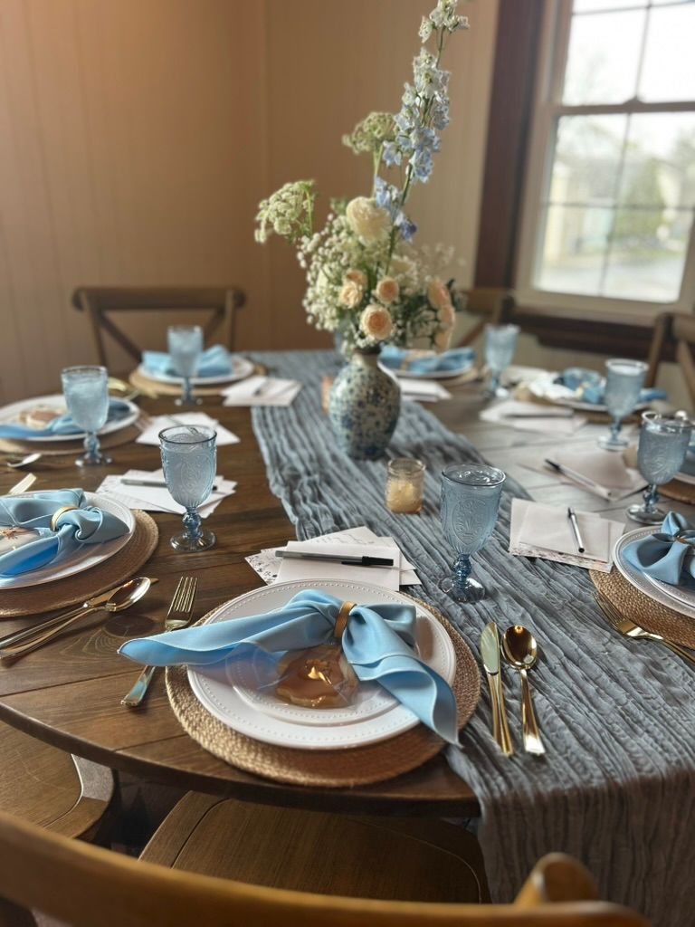 Blue-themed round table setting with ruffled napkins, gold flatware, floral centerpiece, and blue glassware near window.