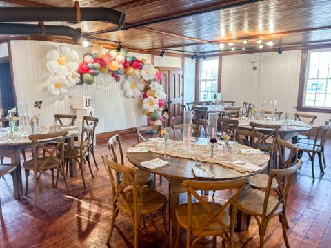Bright event hall with round tables, cross-back chairs, colorful balloon arch, and elegant white table settings.