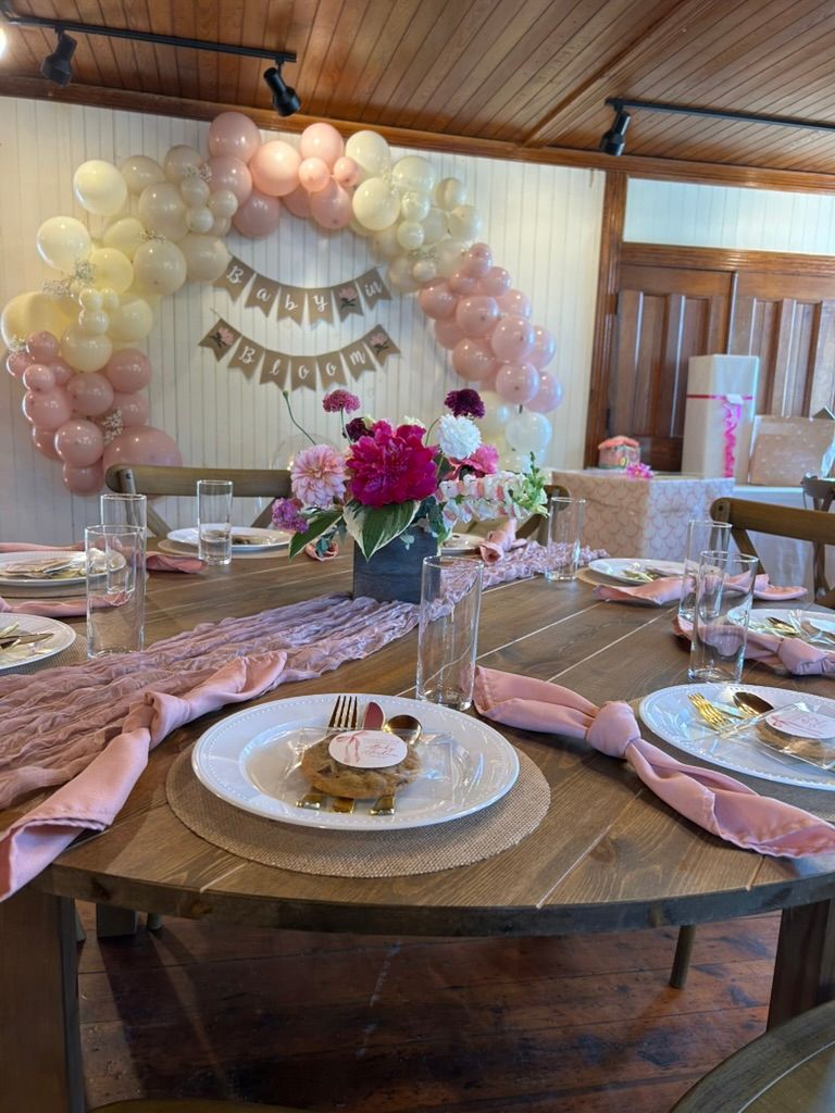 Pink baby shower table with balloon arch backdrop, floral centerpiece, pink napkins, and slice of cake on round table.
