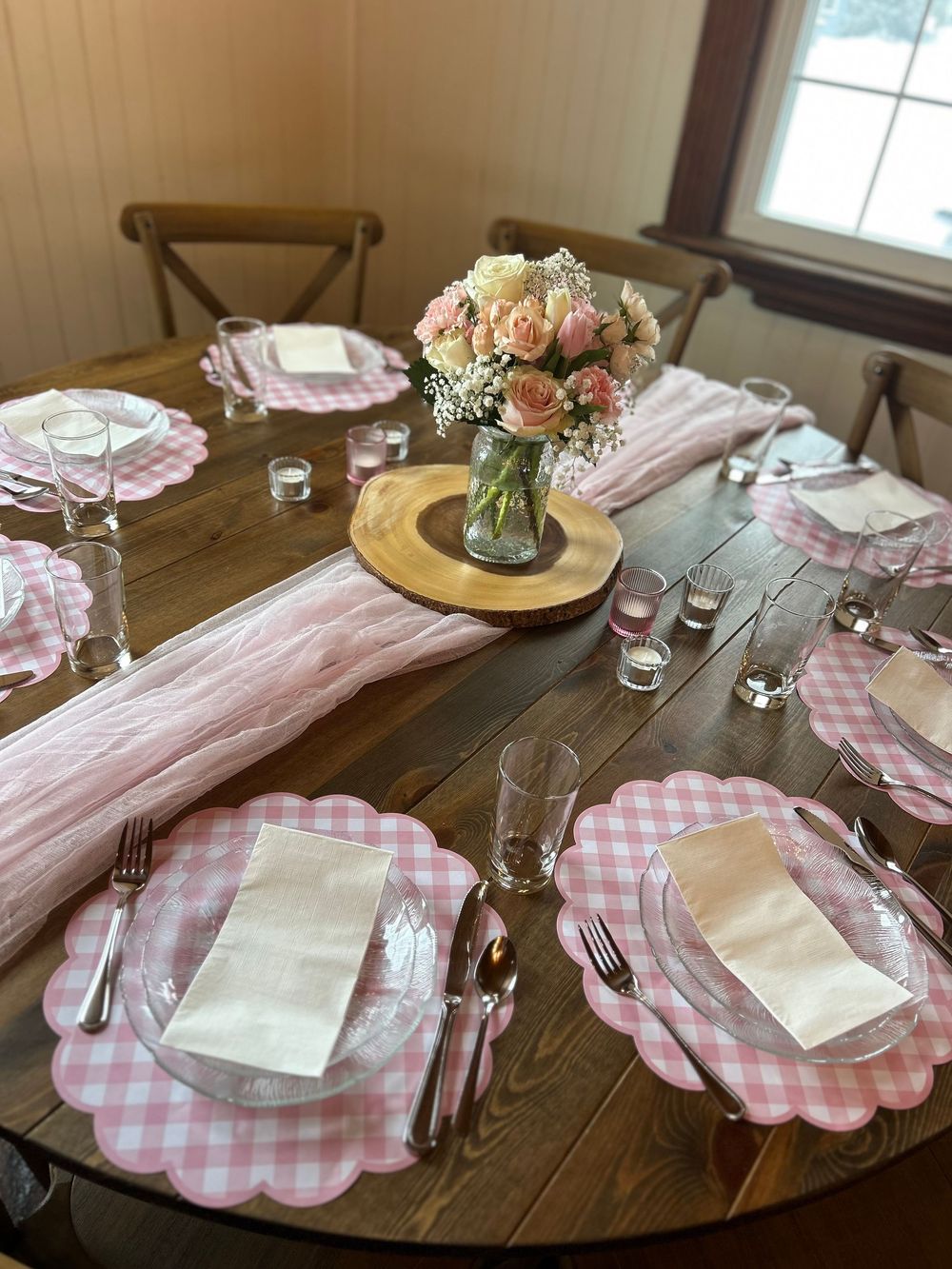 Pink baby shower table with scalloped gingham placemats, pink roses centerpiece, and clear glassware on round wood table.