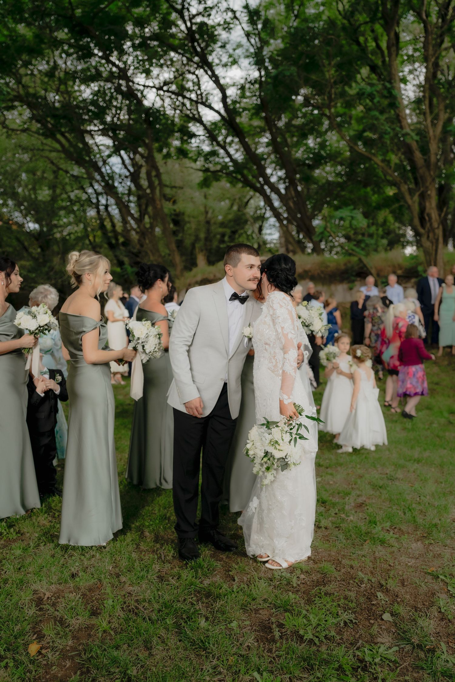 Bride and groom share a kiss outdoors surrounded by bridesmaids in sage green dresses holding white bouquets.