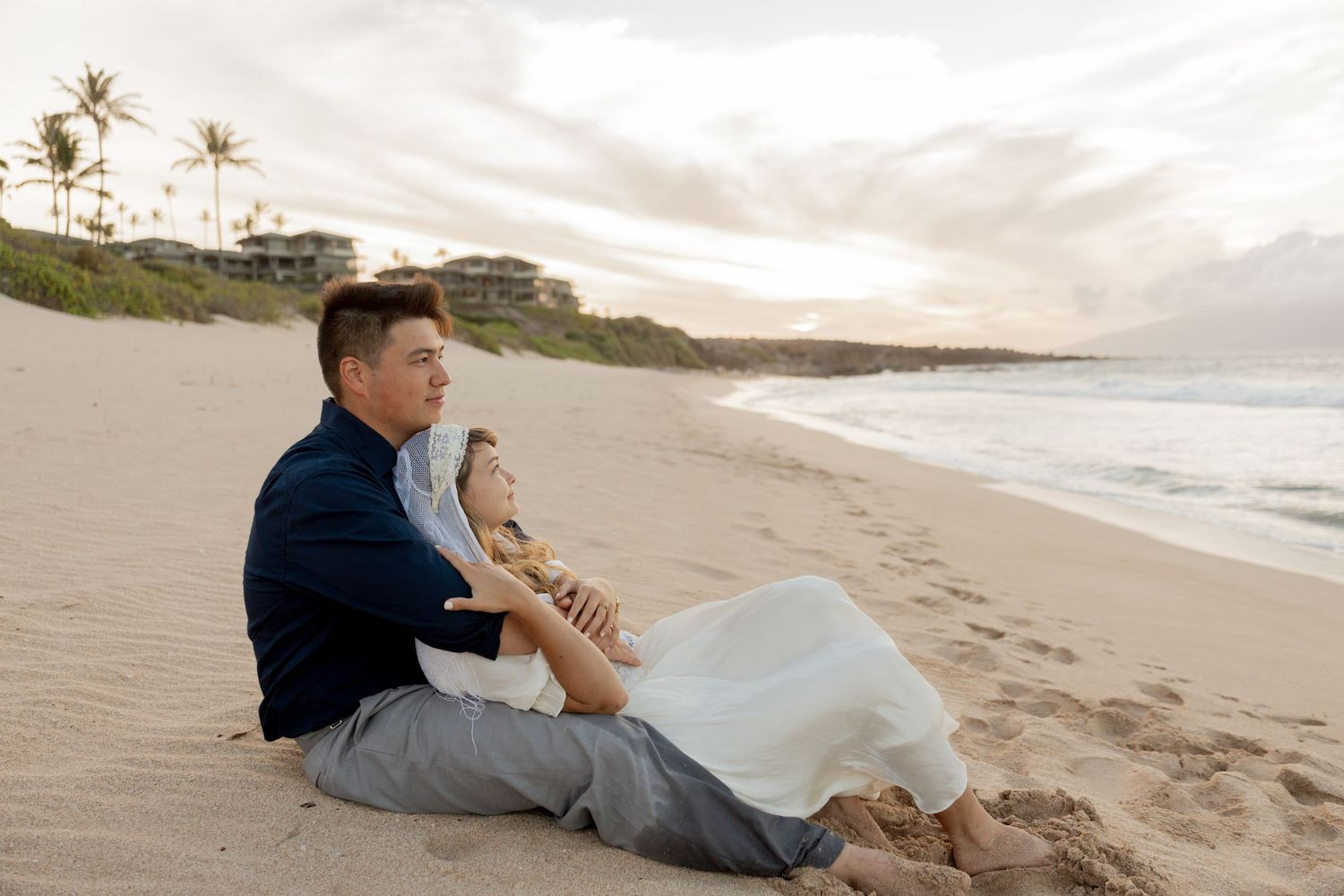 Couple sitting together on a sandy beach at sunset, with ocean waves and palm trees in the background.