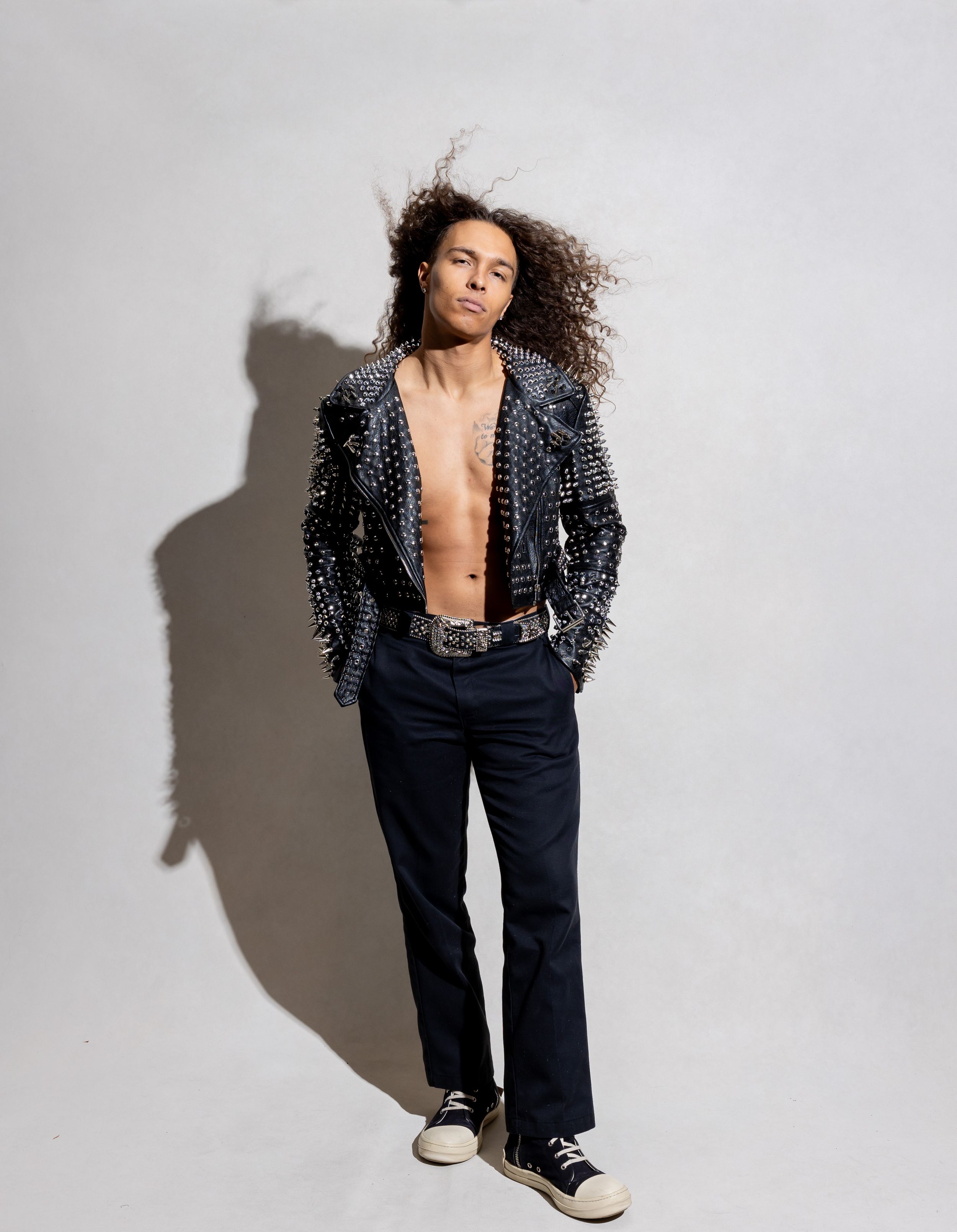 Young man with curly hair wearing a studded leather jacket, open chest, dark trousers, and black sneakers.
