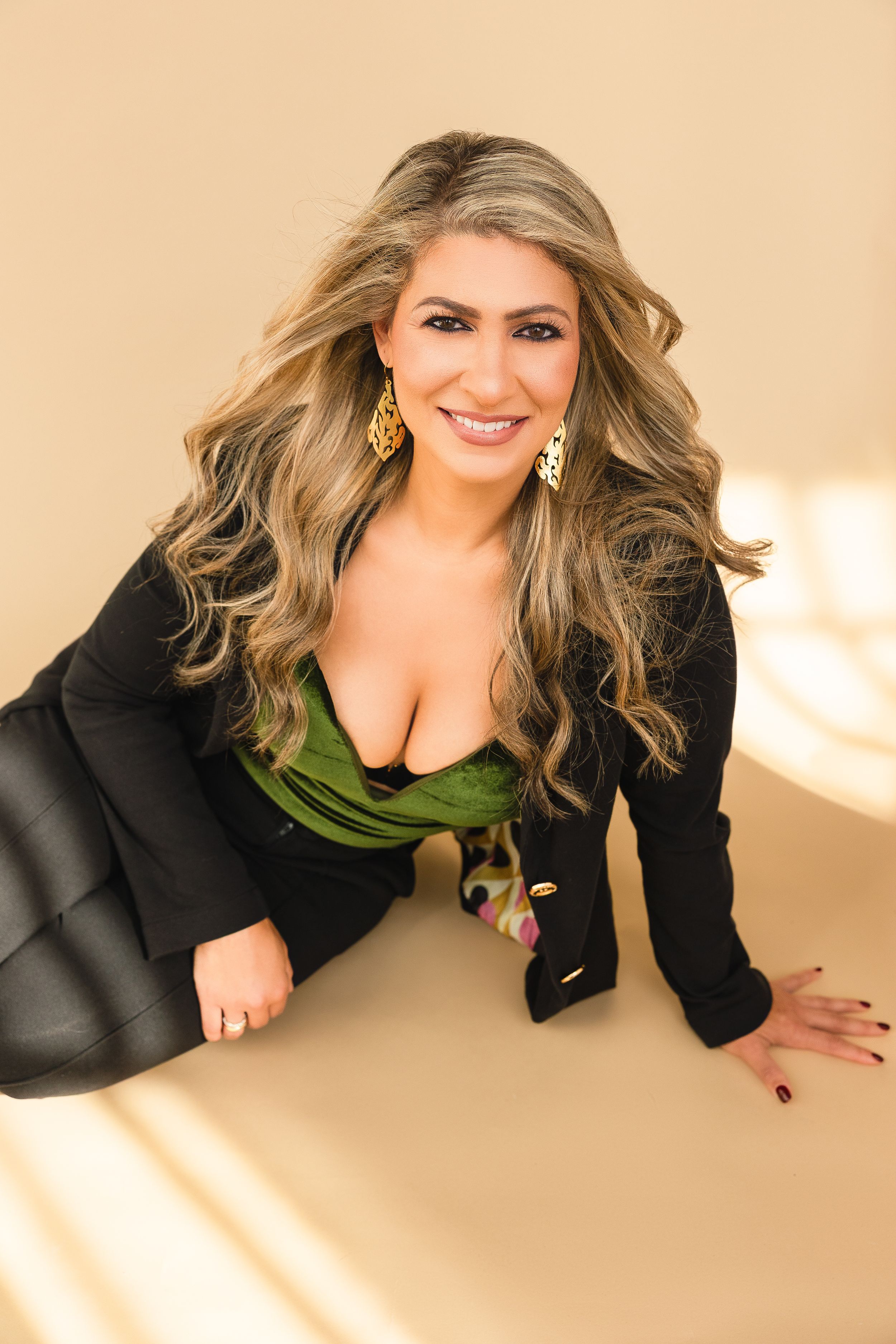 Smiling woman with long blonde hair wearing a black blazer and green top, posing confidently on a beige background.