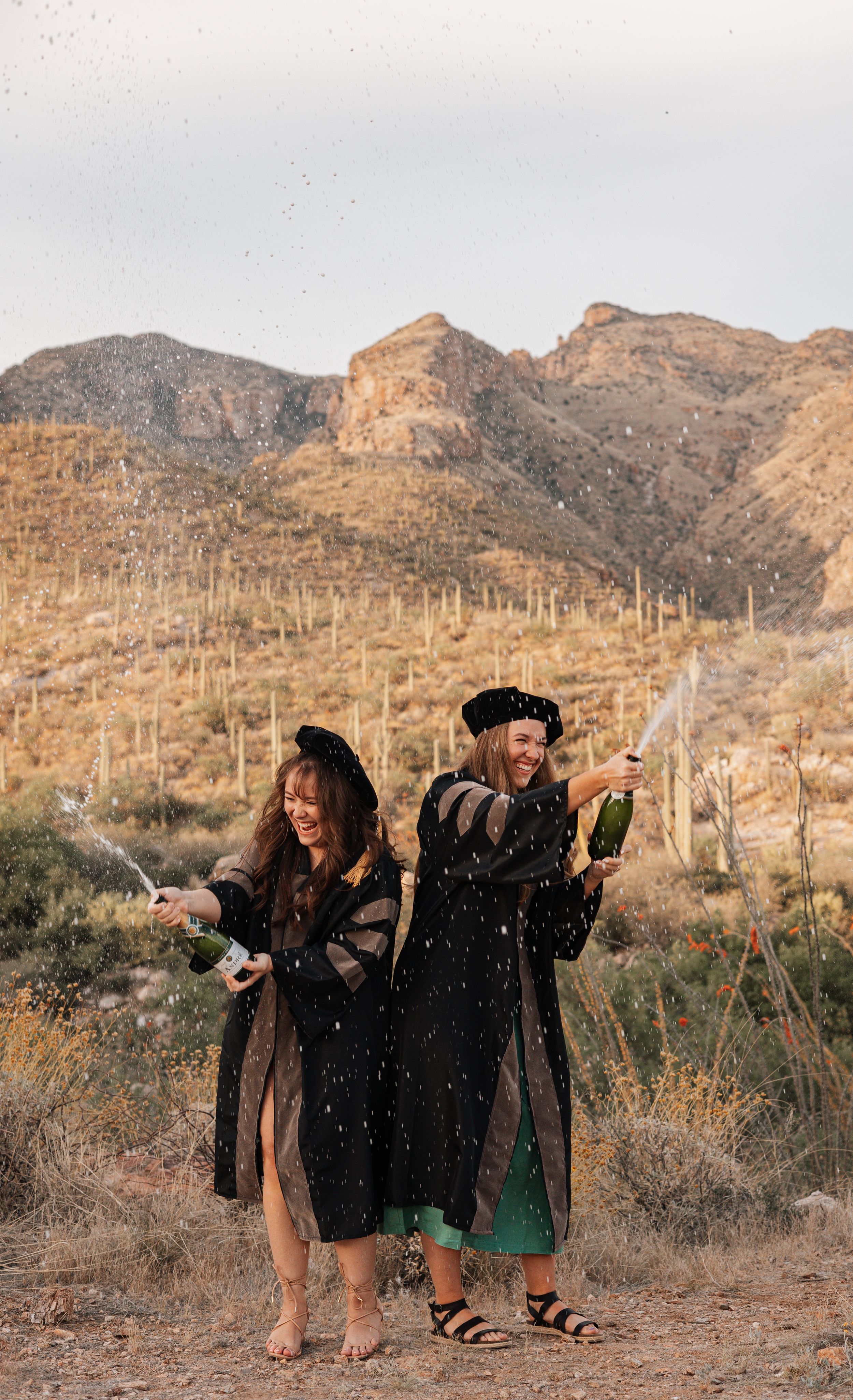Two women in graduation gowns and caps celebrate with champagne in a desert landscape with mountains.