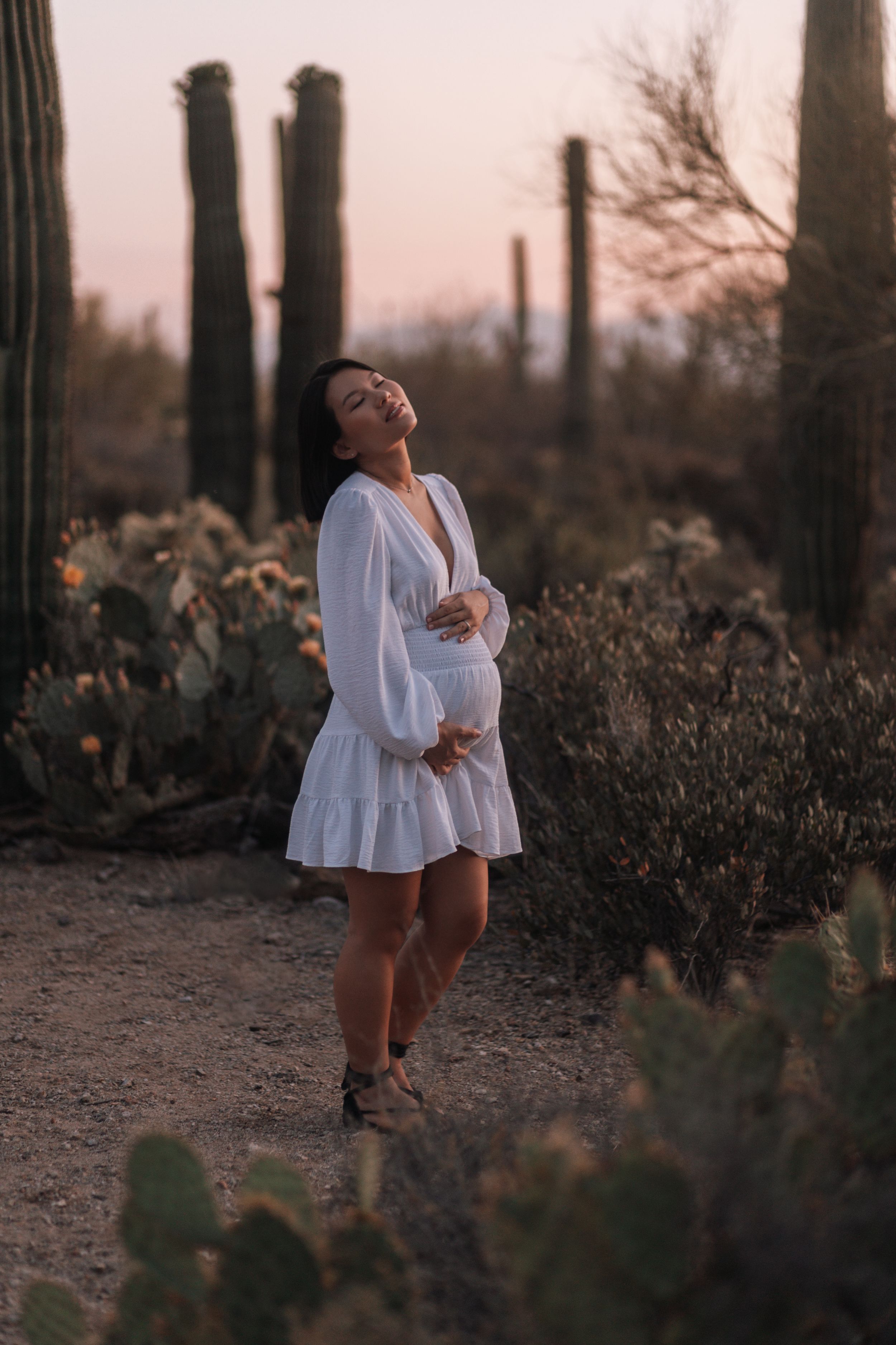 Pregnant woman in white dress cradling baby bump among saguaro cacti at desert sunset.