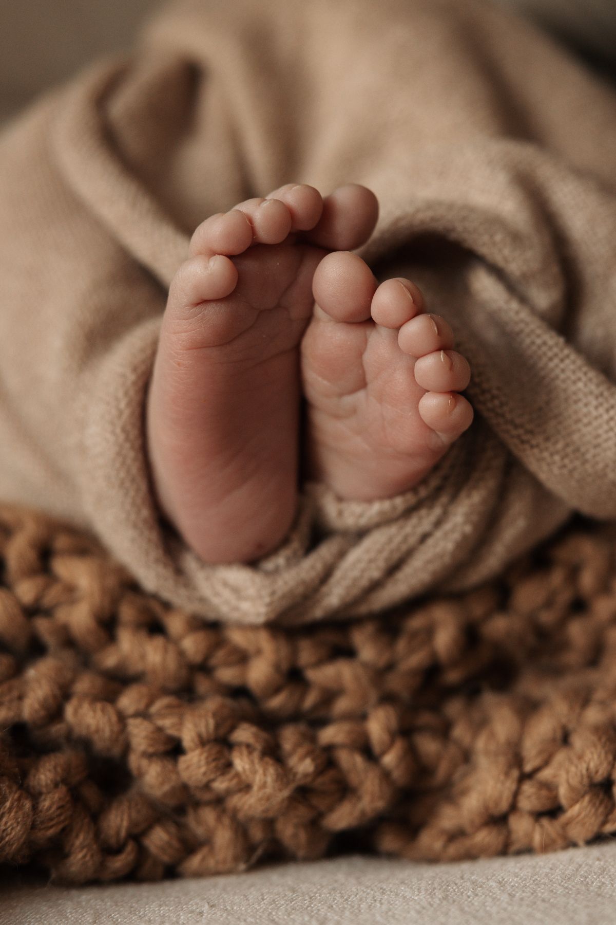 Tiny newborn baby feet peeking out from a soft beige knit blanket on a chunky brown knitted surface.