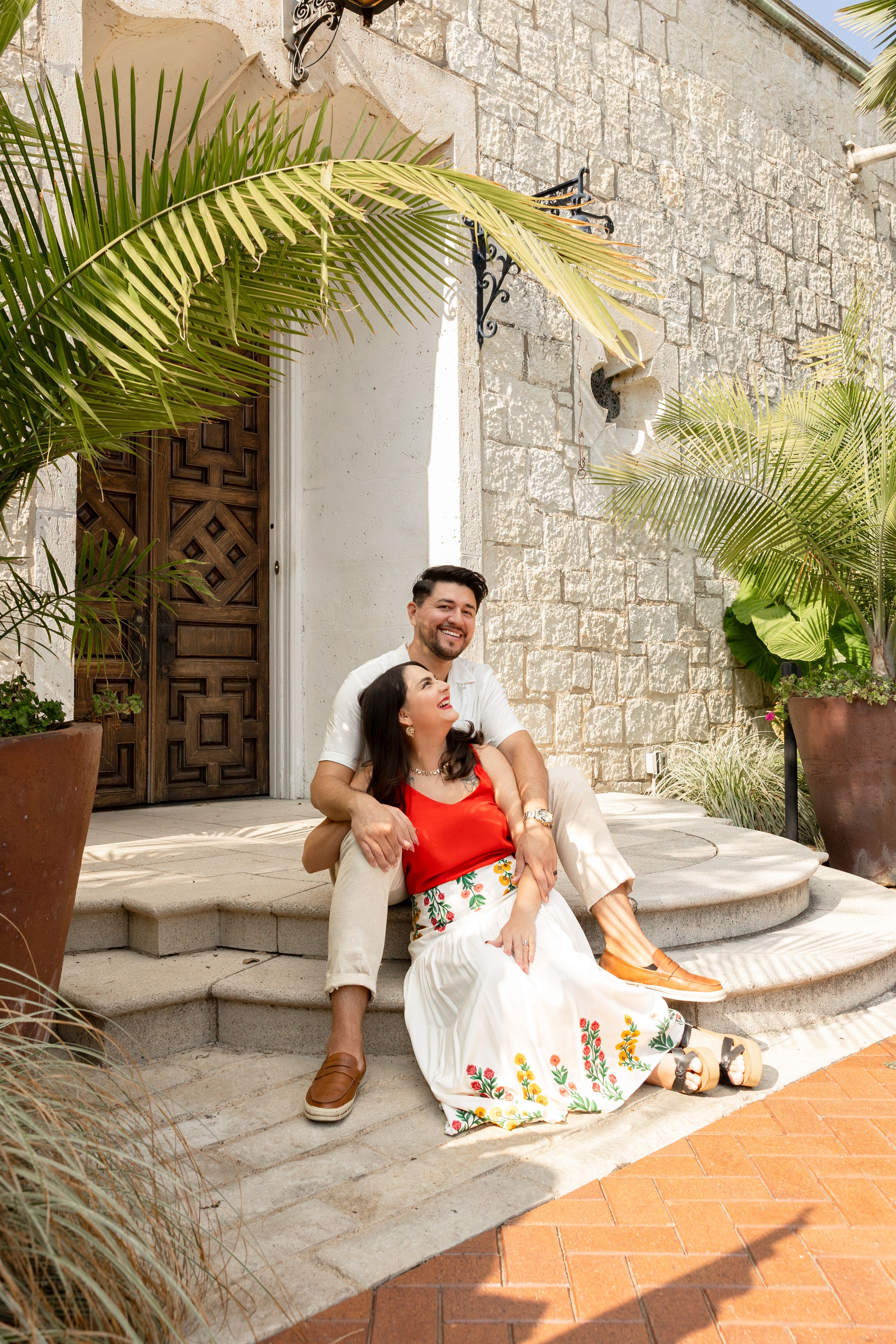 A smiling couple sits on stone steps outside a tropical villa with palm trees and a wooden door.