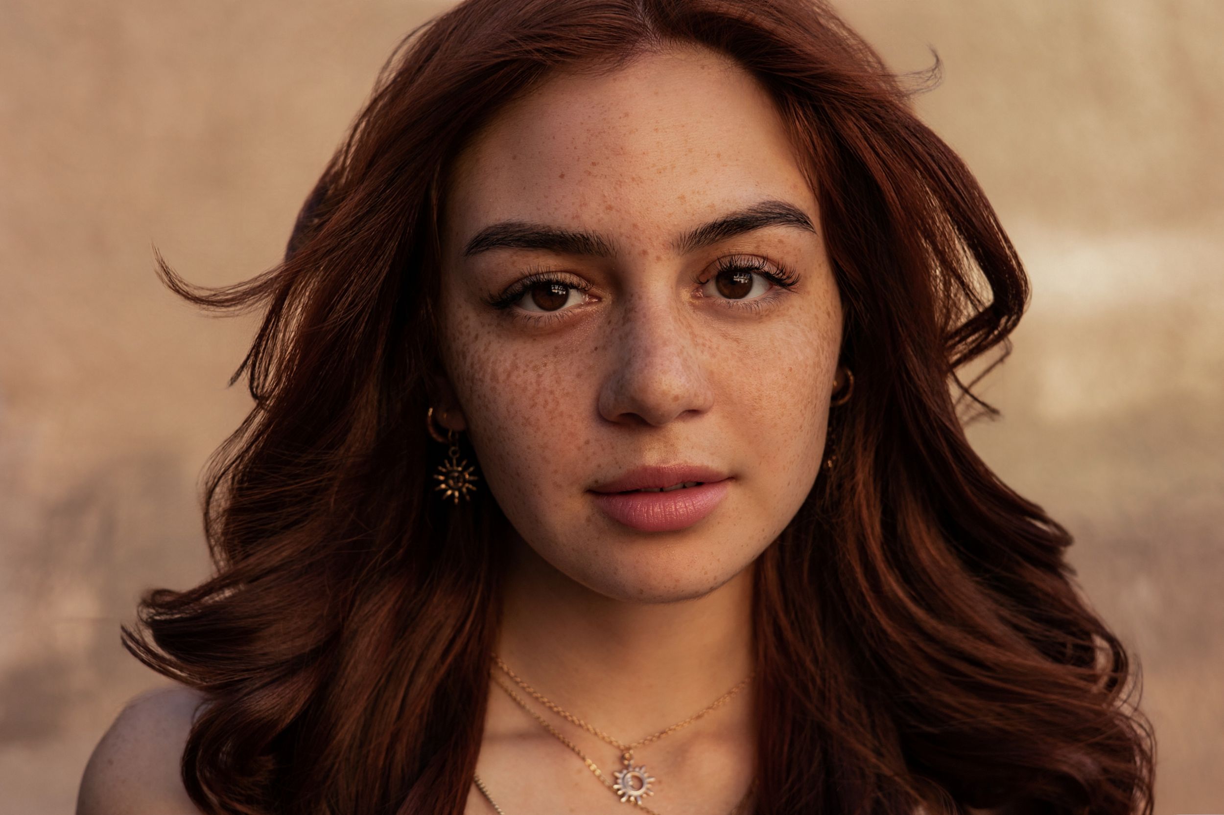 Young woman with auburn wavy hair, freckles, and a necklace poses outdoors in natural light.