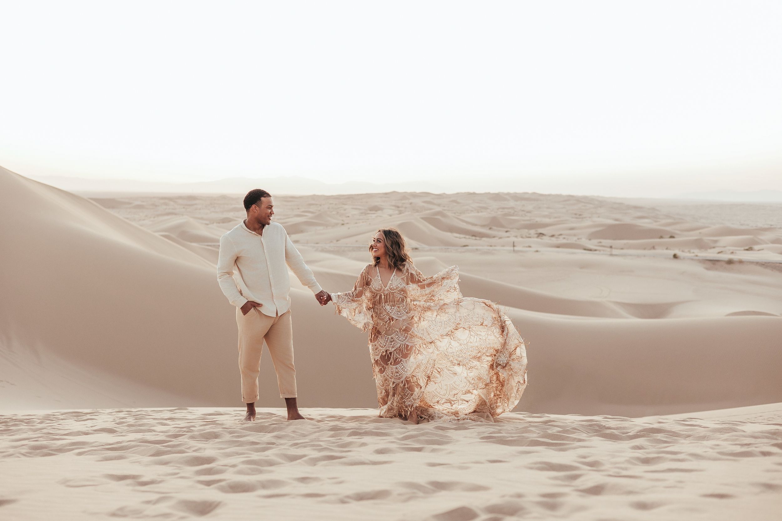 Couple holding hands walking on golden sand dunes in the desert, woman wearing flowing floral gown.