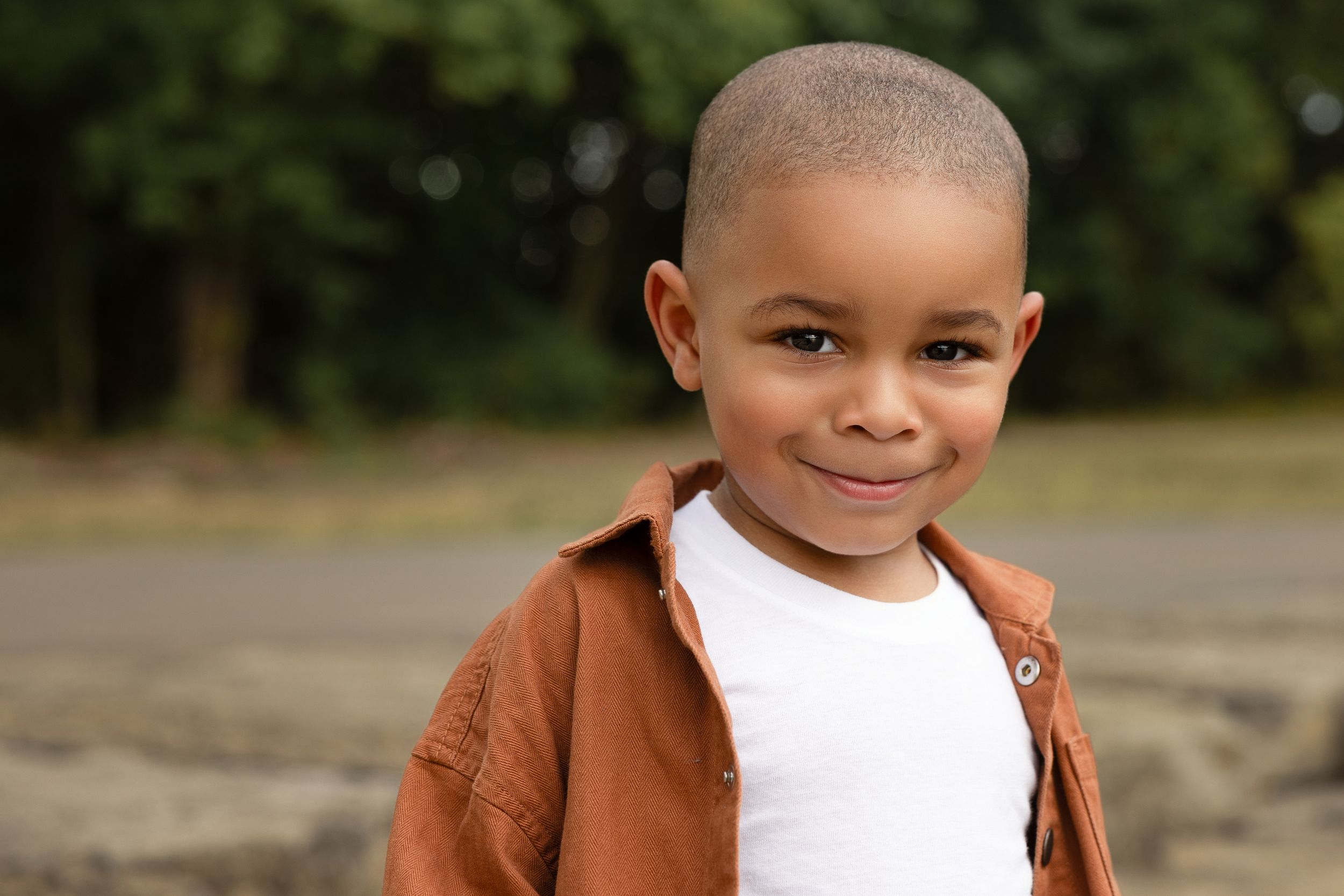 A smiling young Black boy with a shaved head wearing a brown shirt over a white tee, standing outdoors.