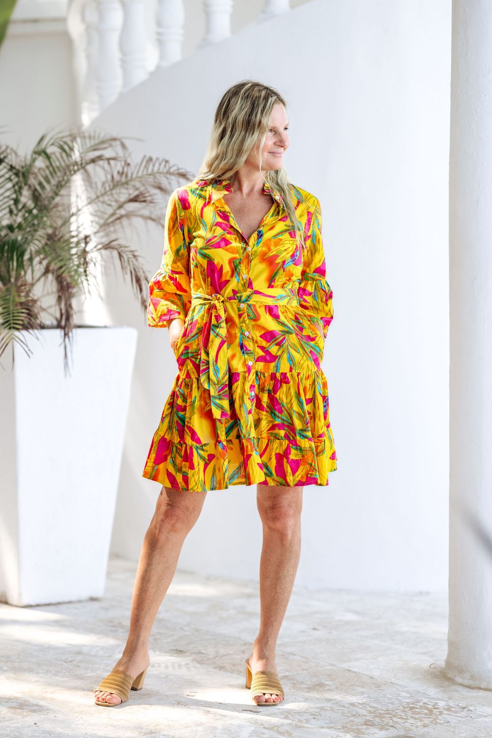 Woman wearing a vibrant yellow tropical print belted shirt dress with ruffle hem and tan mule sandals.