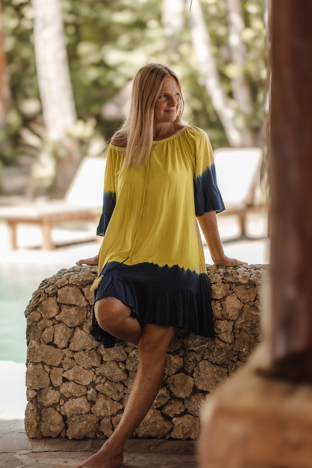 Woman in yellow and navy tie-dye dress sitting on stone wall near tropical pool.