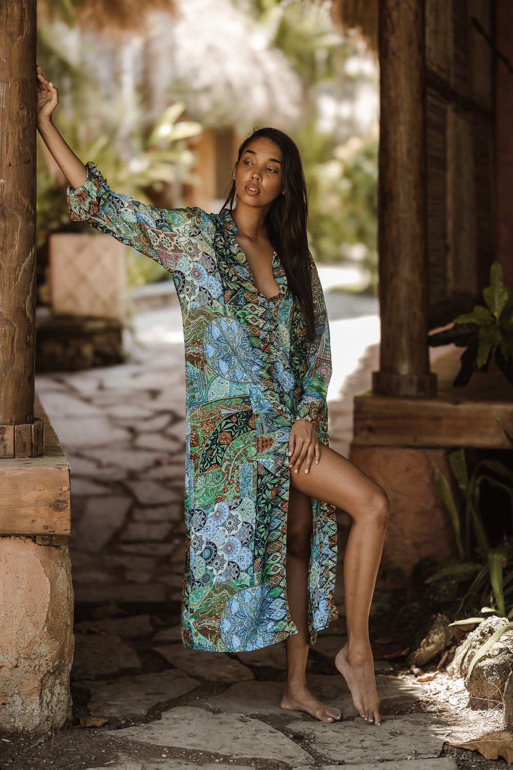 Woman in floral kimono cover-up poses barefoot against rustic wooden pillar in tropical outdoor setting.