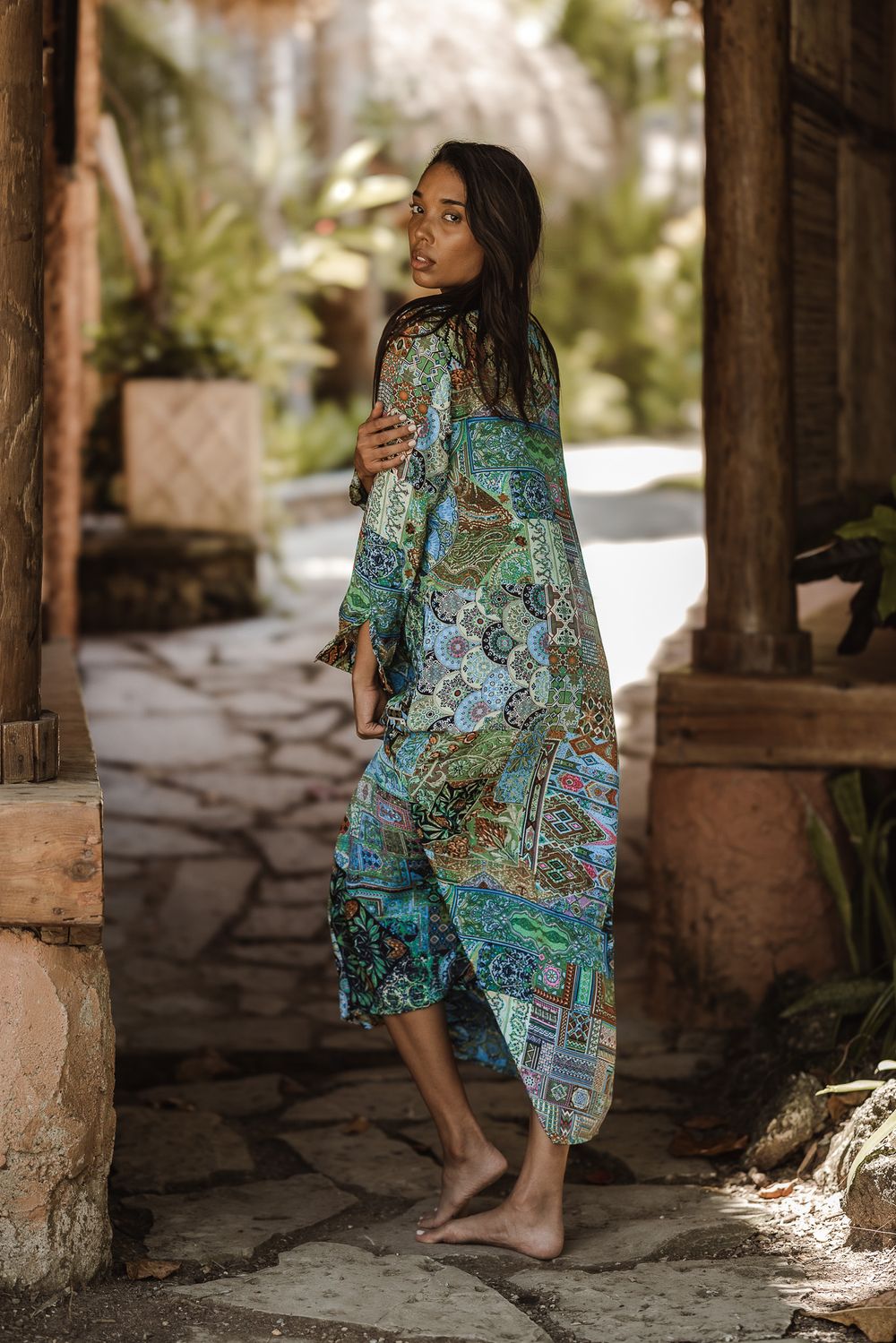Woman in colorful boho patchwork kimono dress standing barefoot on stone path in tropical garden setting.