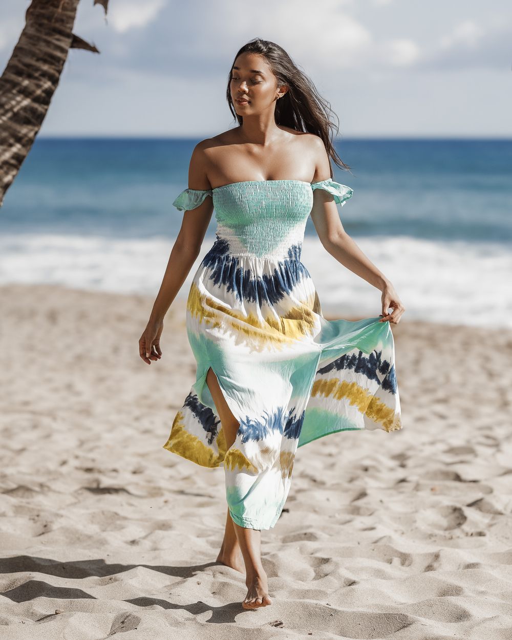Woman in tie-dye off-shoulder maxi dress walking barefoot on sandy beach with ocean waves in background.
