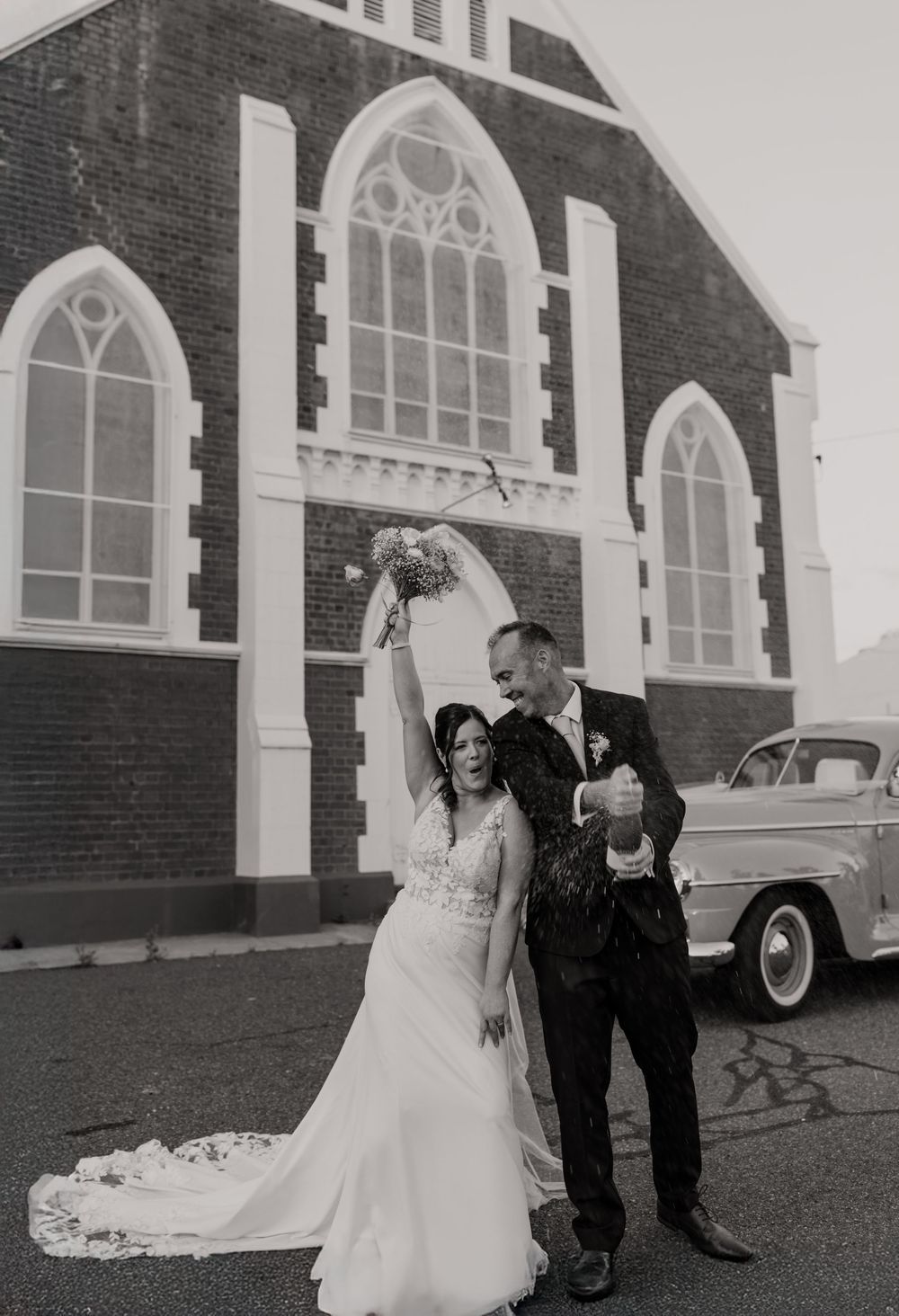Black and white photo of joyful newlyweds celebrating outside a brick church, bride raising bouquet, groom popping champagne.
