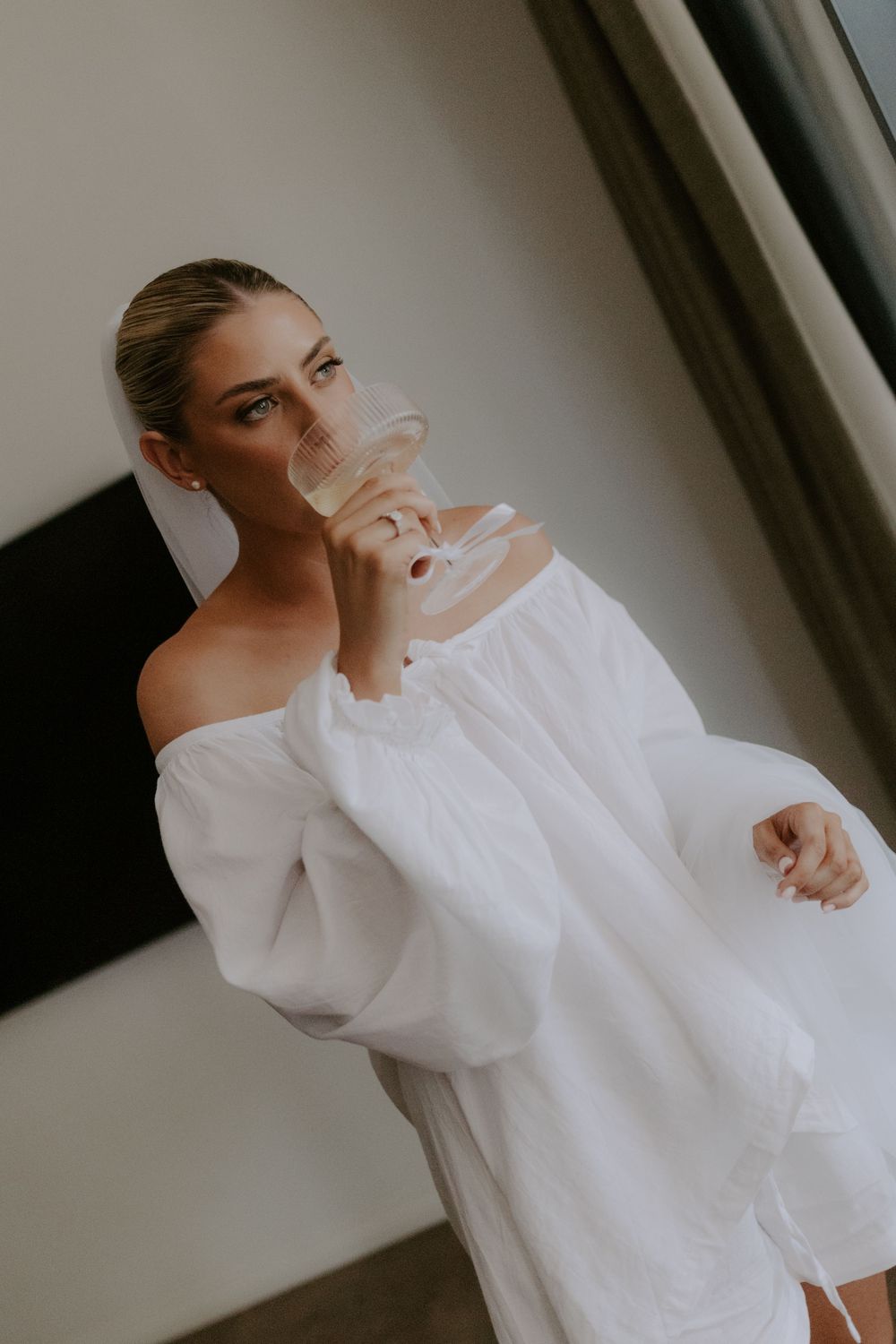Woman in white off-shoulder dress sipping champagne from a glass, elegant bridal style portrait.