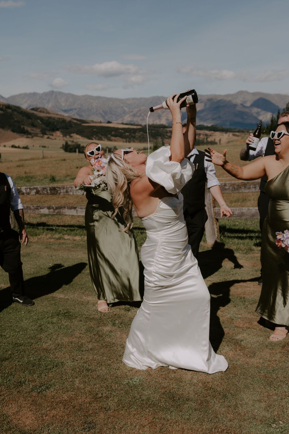 Bride in white satin gown drinking from bottle with bridesmaids in olive dresses, mountain landscape backdrop.