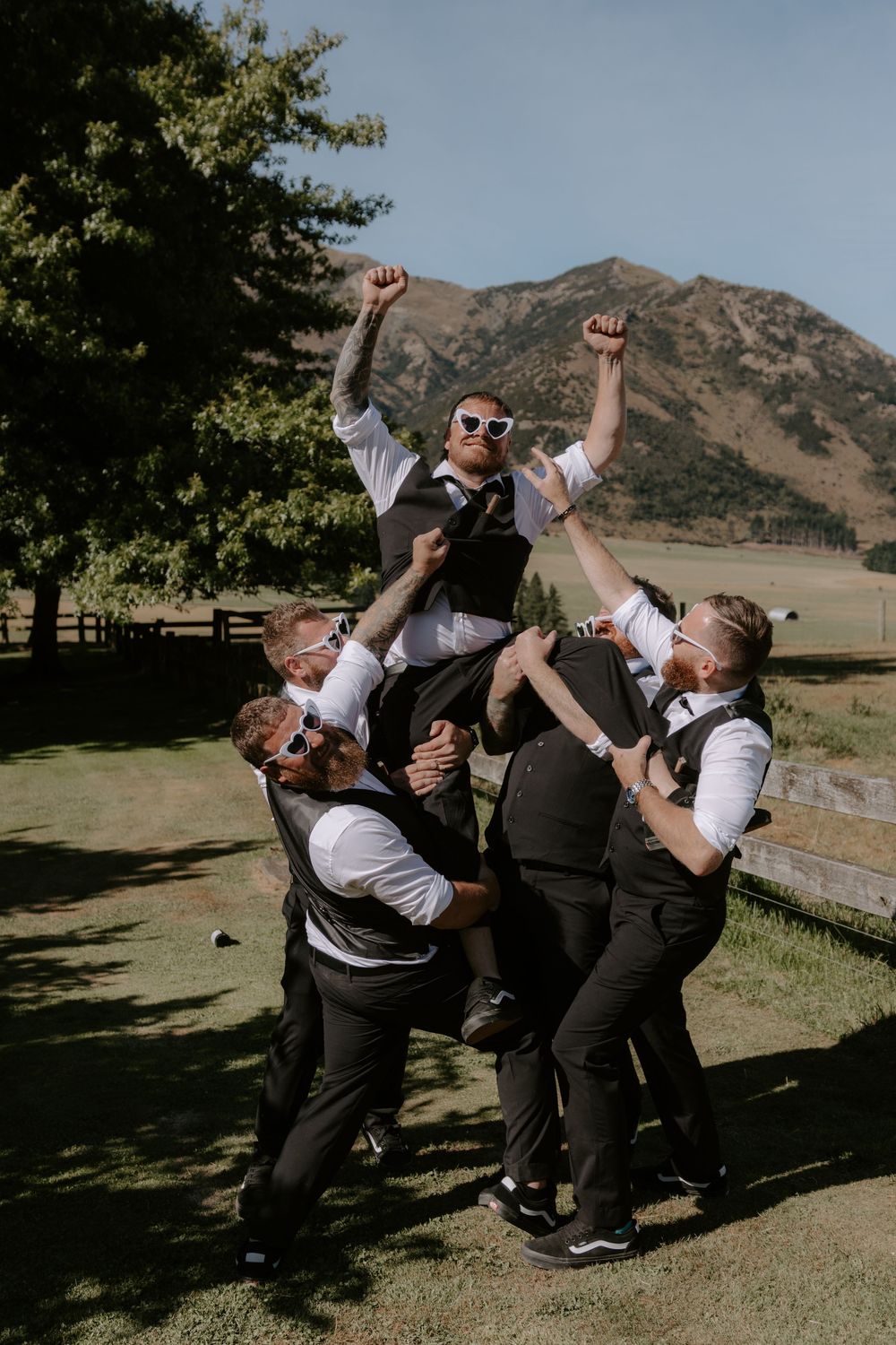 Groomsmen lifting groom in celebration outdoors with mountain backdrop on sunny wedding day in New Zealand.