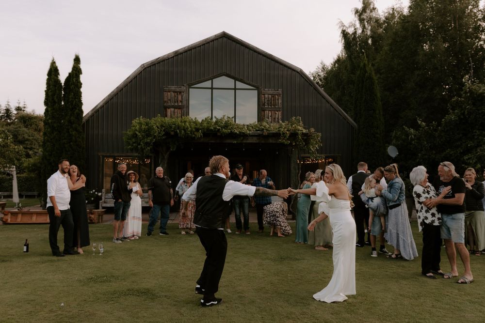 Bride and groom share first dance on lawn outside rustic black barn wedding venue with guests watching.