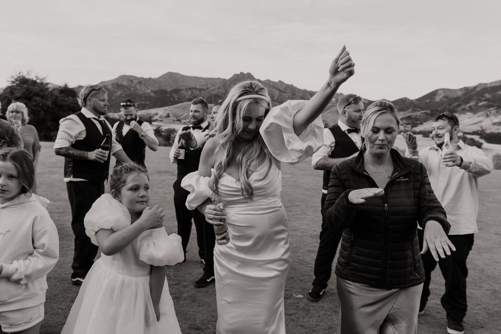 Bride in white gown dances joyfully with guests outdoors, mountains in background, black and white wedding photo.