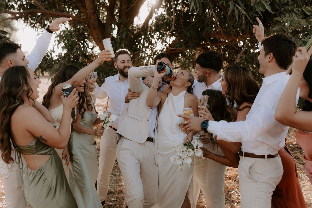 Wedding party celebrates outdoors under trees, bridesmaids in sage green dresses raising glasses with grooms in white suits.
