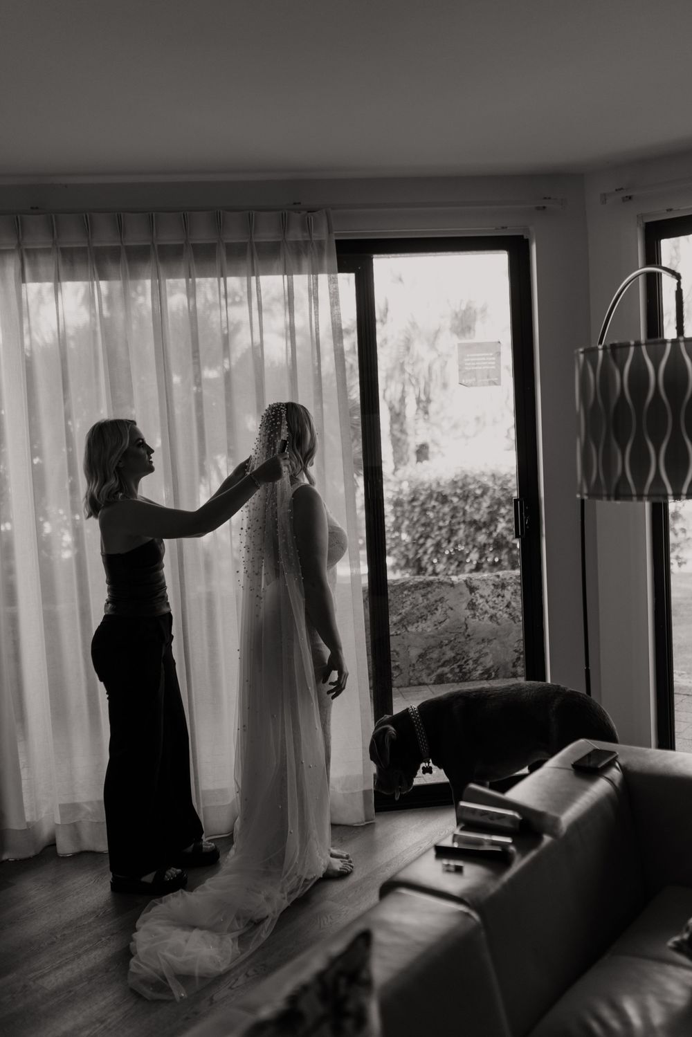 Black and white photo of a bridesmaid helping a bride with her veil near a sunlit window in an elegant room.