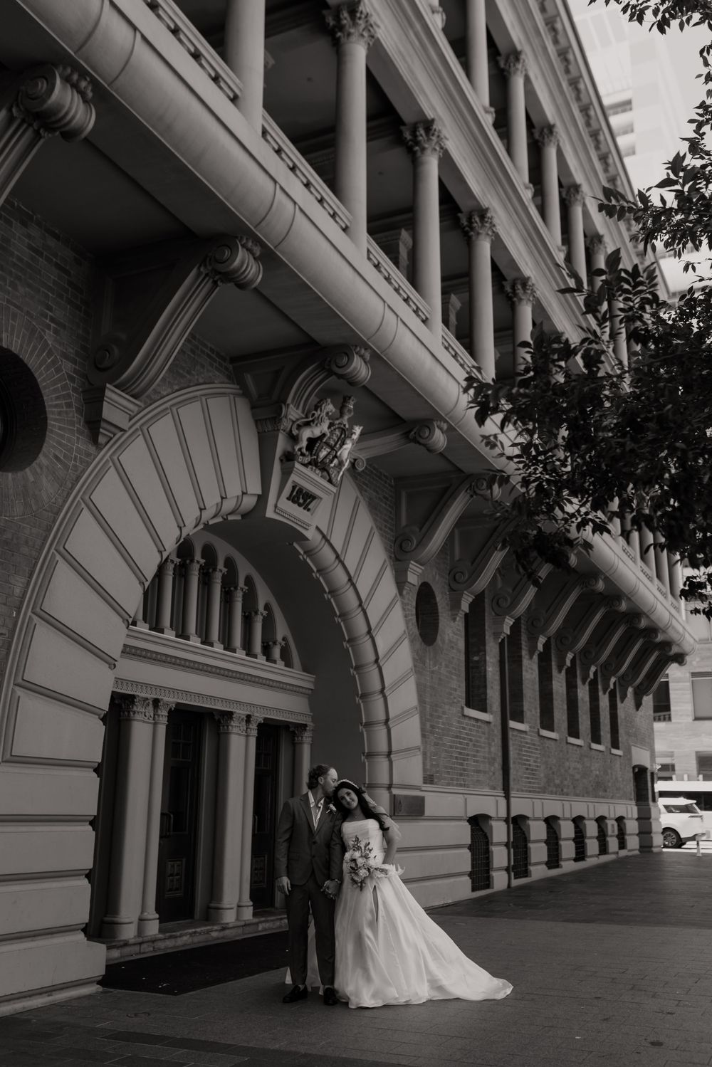 Black and white wedding photo of bride and groom standing under ornate arch of historic building.