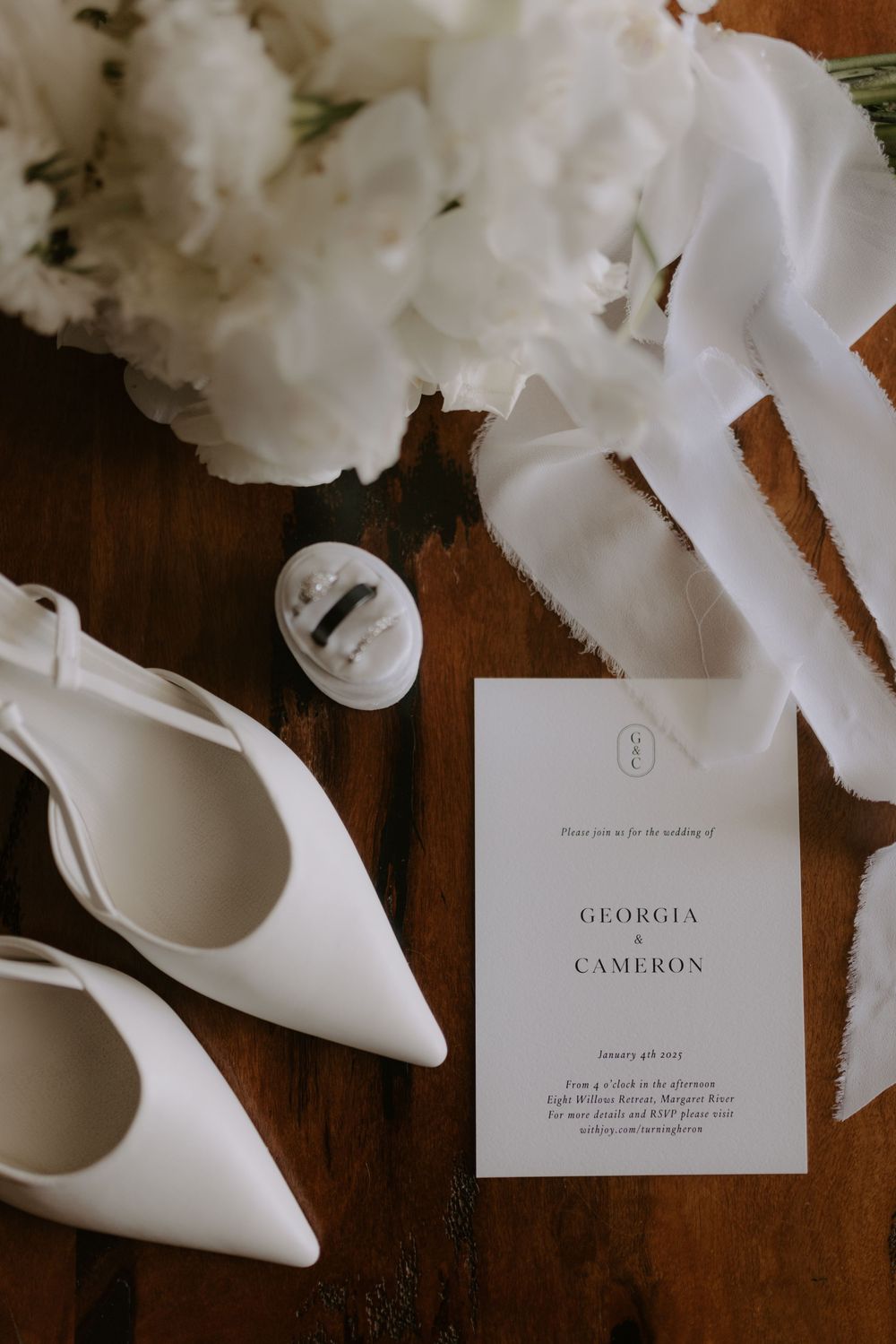 Elegant wedding flat lay featuring white heels, floral bouquet, ring box, and Georgia & Cameron wedding invitation on wood.