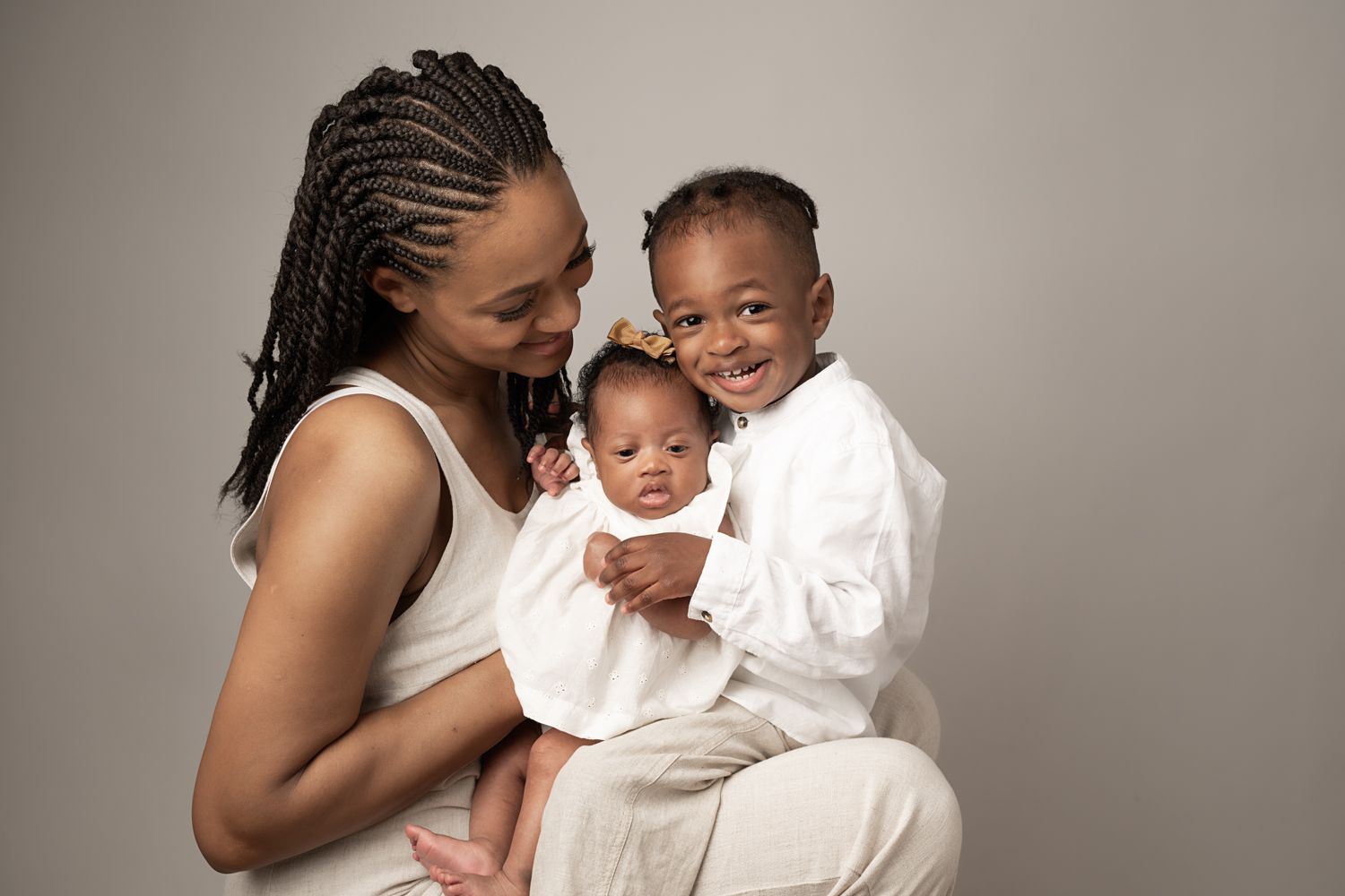 A mother lovingly holds her newborn and toddler, all dressed in white, smiling warmly together.